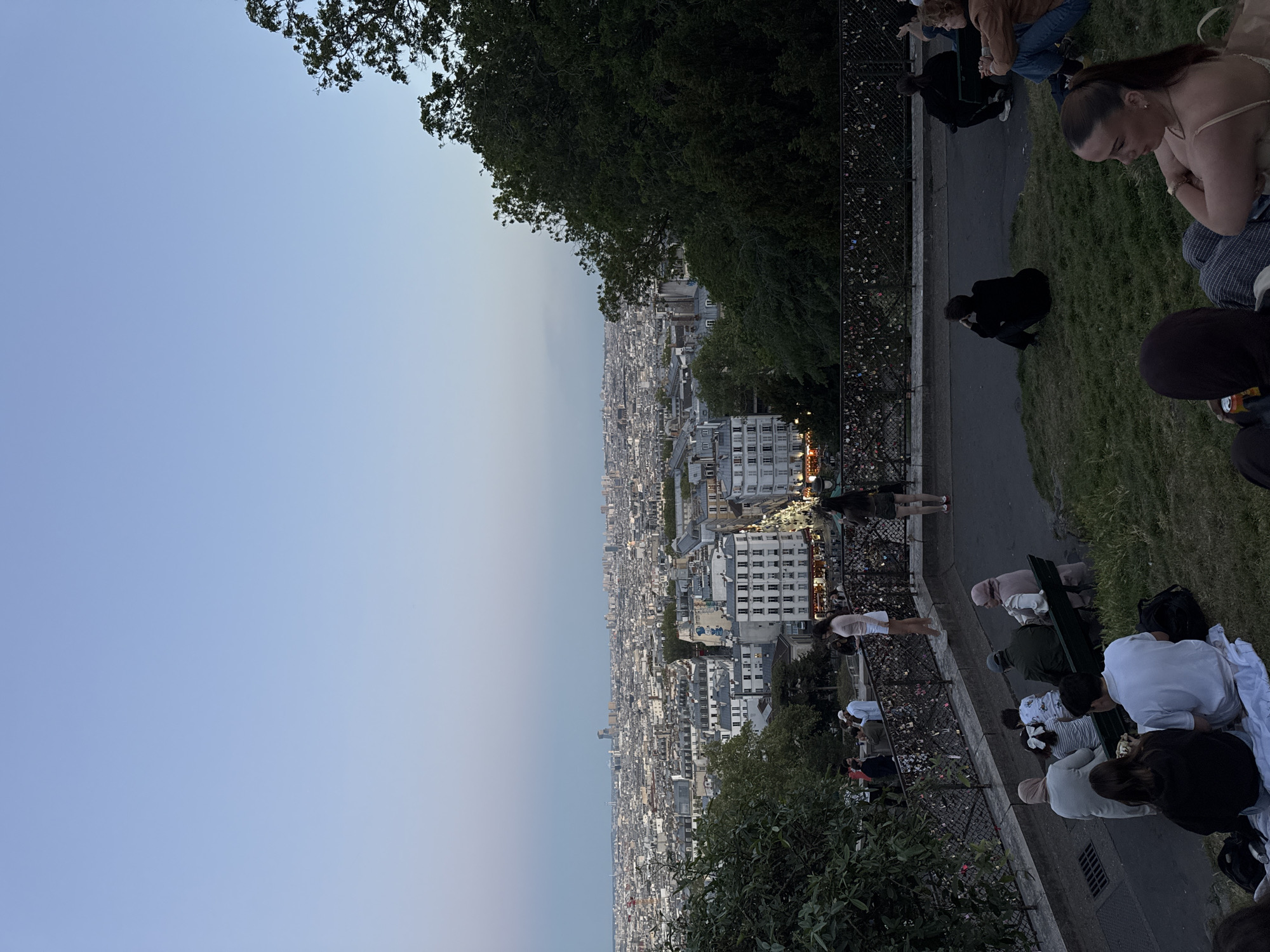 The image presents an elevated, panoramic view of a vast city at dusk, likely Paris, France, given the typical architecture and the presence of "love locks." In the foreground, numerous people are gathered on a grassy slope and a paved pathway, mostly facing away from the camera and looking out over the urban landscape. Some individuals are seated on the grass, while others stand or sit near a dark metal fence that runs along the edge of the viewing area. This fence is visibly adorned with many small "love locks."
To the left, green foliage and tree branches partially frame the scene, contrasting with the expansive city beyond. The cityscape itself stretches into the horizon, characterized by dense clusters of buildings with varied heights and typical European architecture, including numerous apartment blocks and rooftops. As it is early evening, the soft, ambient light suggests dusk; the sky transitions from a light, hazy blue near the horizon to a deeper blue overhead, while faint city lights are beginning to twinkle in the distance. The overall atmosphere appears tranquil, with people enjoying the extensive view as day transitions to night. No visible text is present in the image.