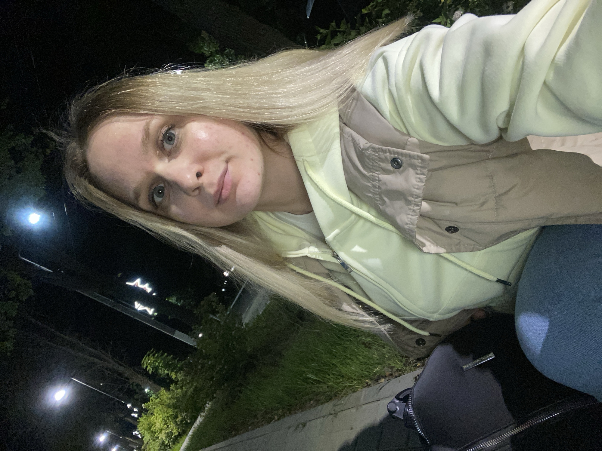 A young woman with blonde hair takes a selfie outdoors at night. She is wearing a pale yellow hoodie and a tan vest. The background features streetlights illuminating trees and foliage, suggesting a park or a street in a city. A black bag is visible to her left, and she is seated next to a paved walkway. The lighting indicates artificial illumination from street lamps. The scene appears to be a casual, nighttime setting within a populated area of Chaikovsky, Russia.