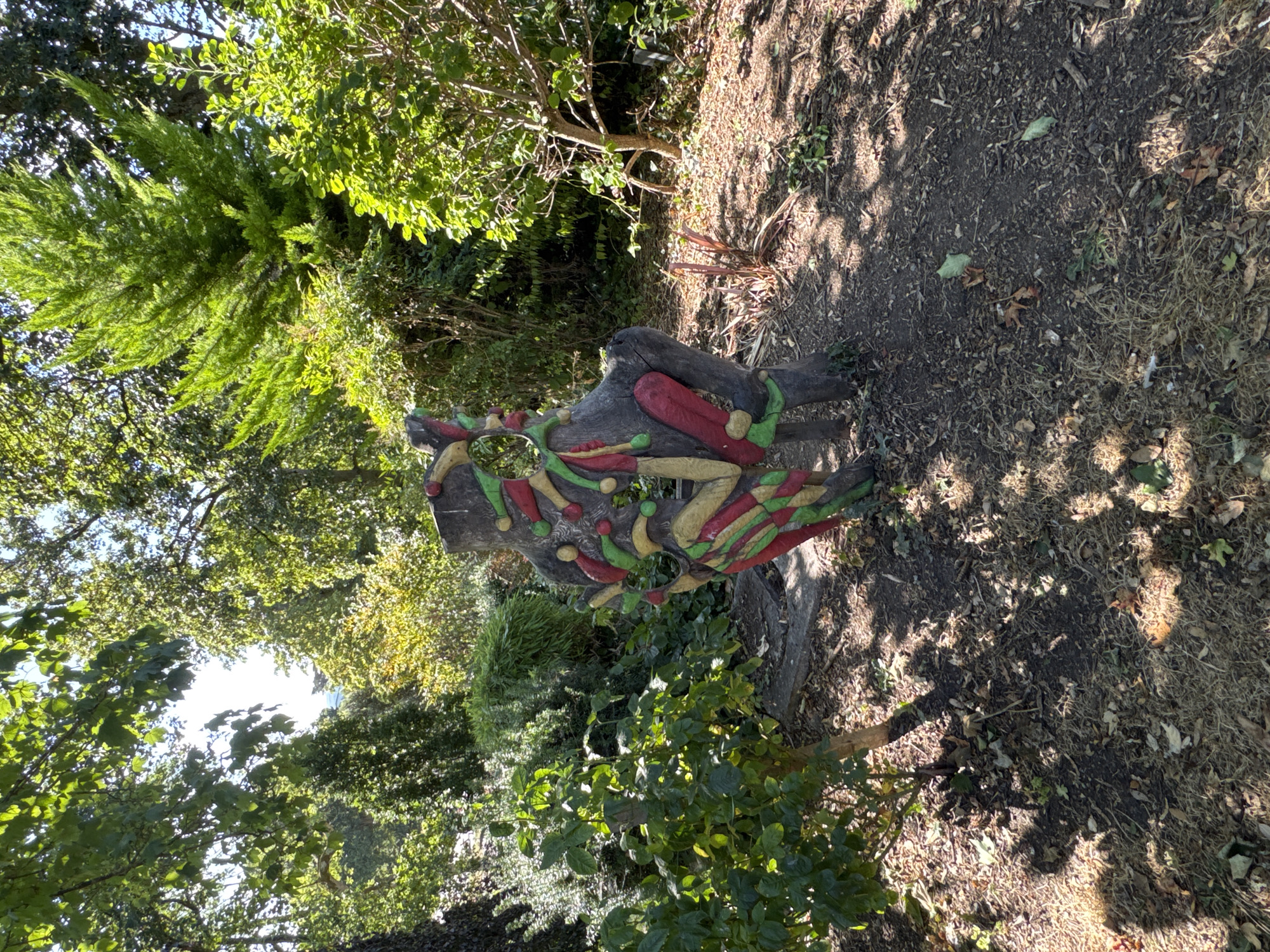 The image depicts a whimsical sculpture made from a tree stump, decorated with colorful, organic shapes in red, yellow, and green. The sculpture is situated in an outdoor setting, surrounded by lush green foliage, trees, and dappled sunlight. The ground is covered with dry leaves and mulch. The scene suggests a garden or park environment. This sculpture is located in the grounds of Cahir Castle in Cahir, Ireland.