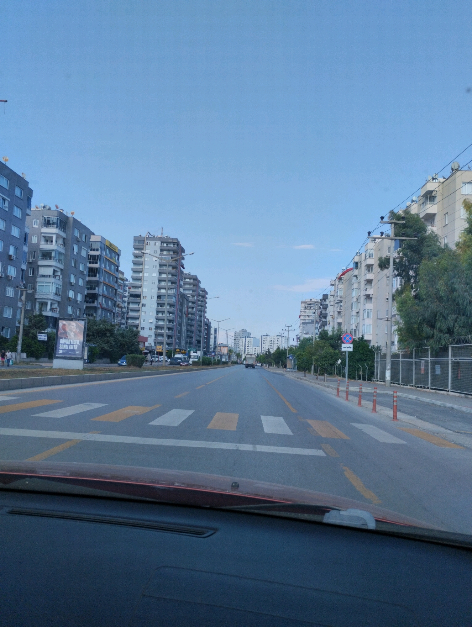 The image shows a street scene in Mezitli, Turkey, looking down a wide avenue lined with multi-story apartment buildings on both sides. The sky is clear and blue. The road has multiple lanes and a pedestrian crossing with yellow and white markings. There are cars on the road and a truck in the distance. Some trees and streetlights are visible. On the left side, a billboard displays a portrait and Turkish text. On the right side, there is a fence and more trees. The photo appears to be taken from a vehicle, as the lower part of the dashboard and windshield are visible.