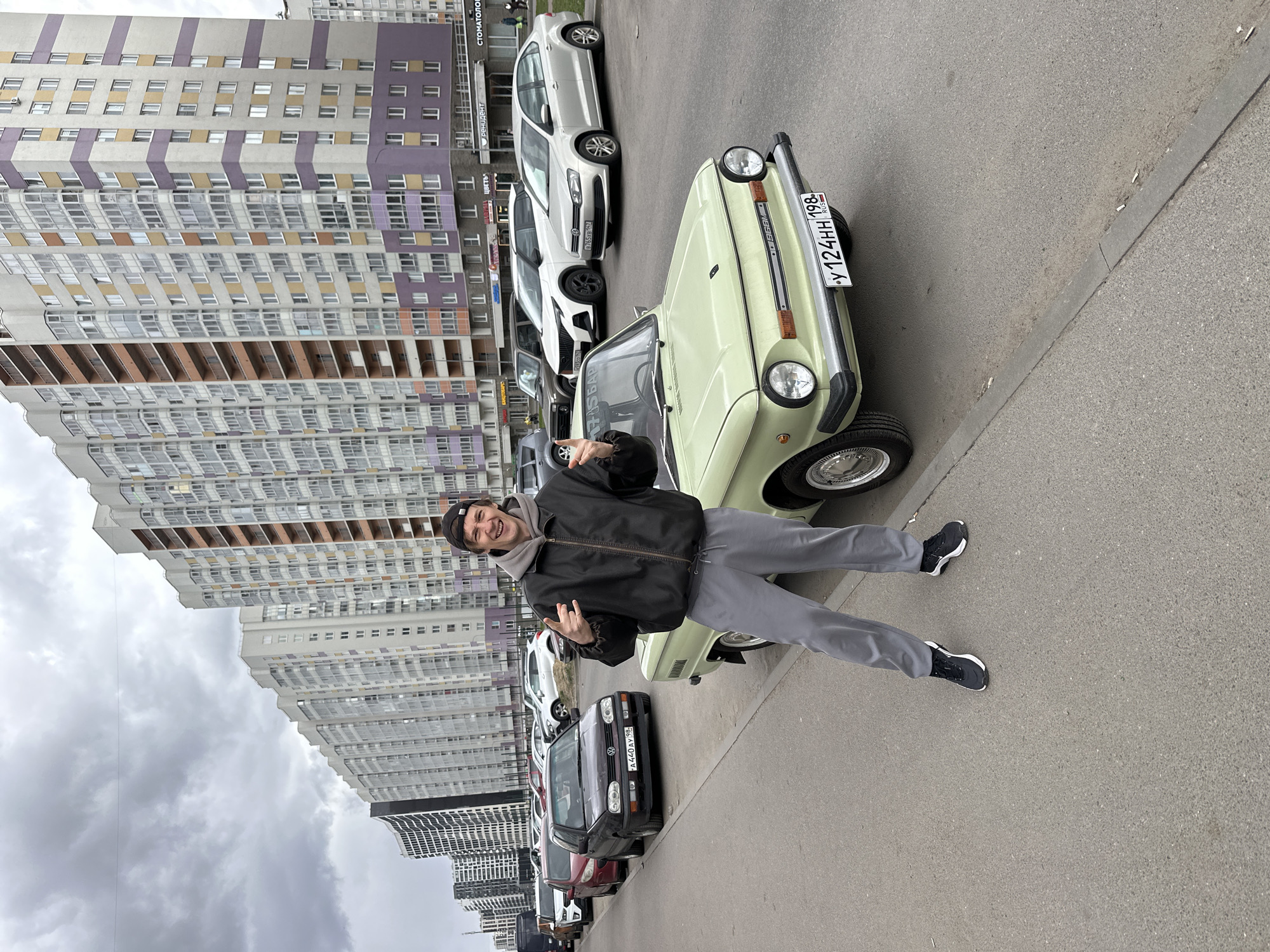 This image, rotated 90 degrees counter-clockwise, depicts a cheerful young man posing next to a light green vintage car on an asphalt street in Murino, Russia. The man, likely in his 20s or 30s, is centered in the frame, wearing a black baseball cap backward, a dark jacket over a light gray hoodie, gray pants, and black sneakers. He displays a wide smile and makes "rock on" hand gestures with both hands, conveying a sense of happiness and excitement.
The vintage car, a compact model, is equipped with a Russian license plate reading "У 124HH 198 RUS". Behind it, a line of modern vehicles is parked along the curb, including a white Volkswagen Polo and a dark Volkswagen Golf with the license plate "A 440AY 98 RUS".
The background is dominated by numerous tall, multi-story residential apartment buildings. These structures feature a distinctive architectural style with horizontal stripes in light gray, purple, and yellow or beige colors. The sky is overcast and cloudy, indicating daytime with diffused light. Visible Cyrillic text on one of the buildings includes "СТОМАТОЛОГИЯ" (Dentistry) and "РЕНТГЕНТ" (X-ray), affirming the urban setting. The overall scene captures a lively moment within a modern residential area.