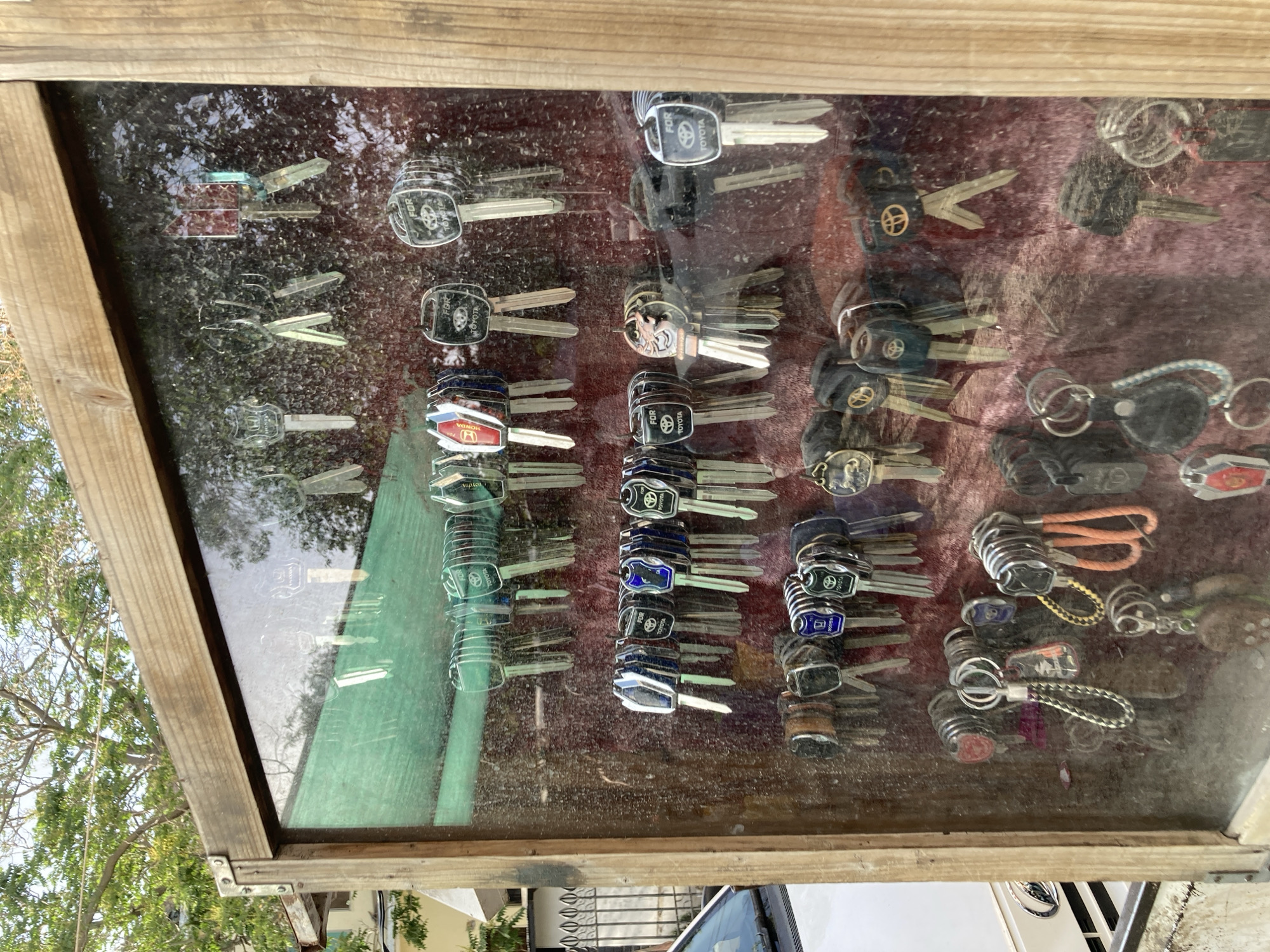 Captured at an oblique angle, the photograph presents a display case, presumably outdoors, housing an array of car keys. The case has a wooden frame, and the keys are arranged against a red and green background. The keys are largely grouped by car brand. Among those keys are Toyota and Honda, with some attached to keychains. The reflection in the glass obscures a clear view, but trees and portions of a building are visible, indicating an outdoor setting in Quetta City Tehsil, Pakistan. The scene is devoid of any human presence.