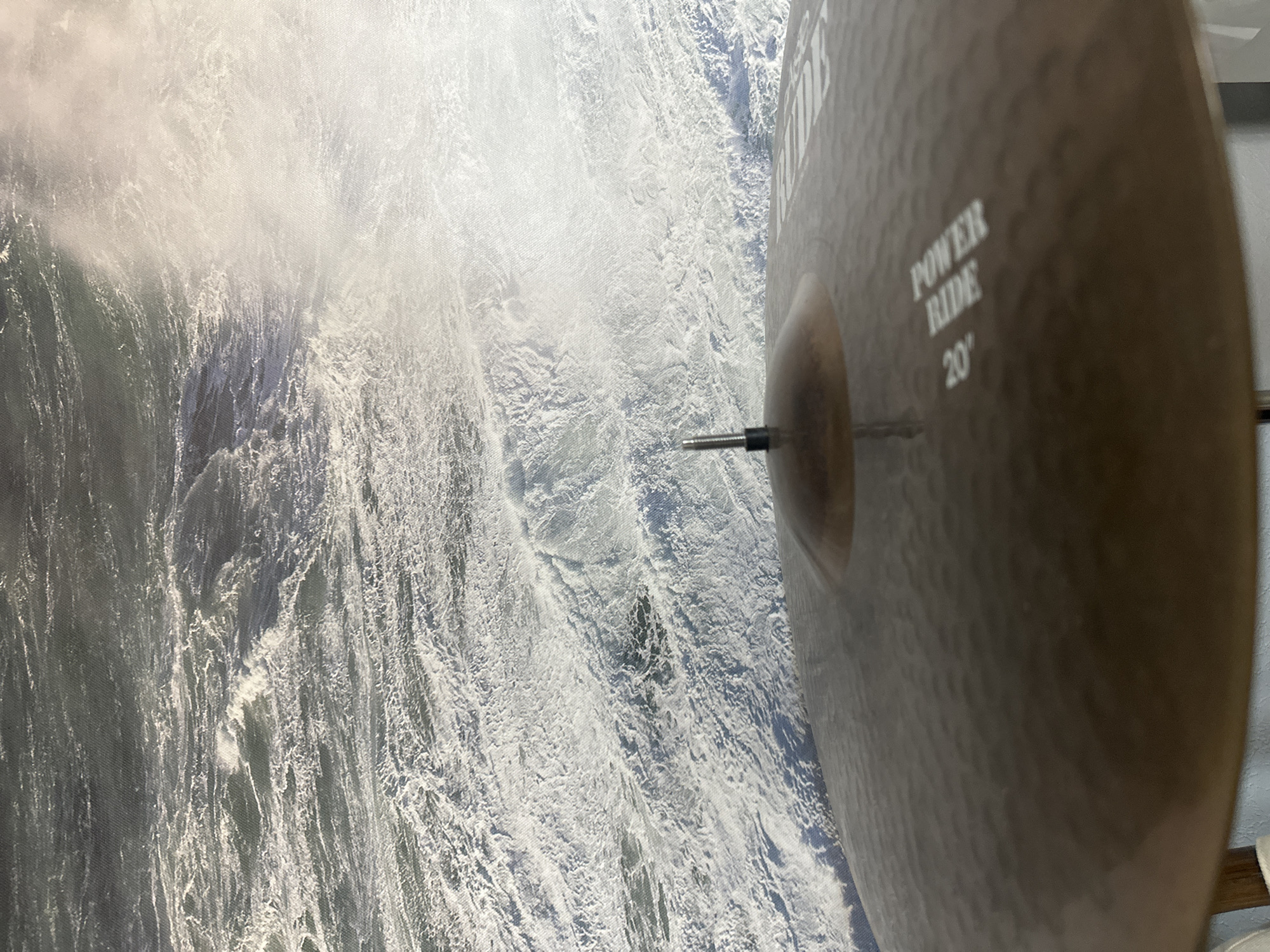 The image shows a close-up view of a cymbal, identified as a "POWER RIDE 20", with a drumstick tip attached to its center. In the background, a turbulent ocean with large waves and spray is visible. The perspective suggests the cymbal is part of a drum kit, possibly placed outdoors near the coast. There is no visible indication of the city of Nevinnomyssk, Russia, or any people present in the scene.