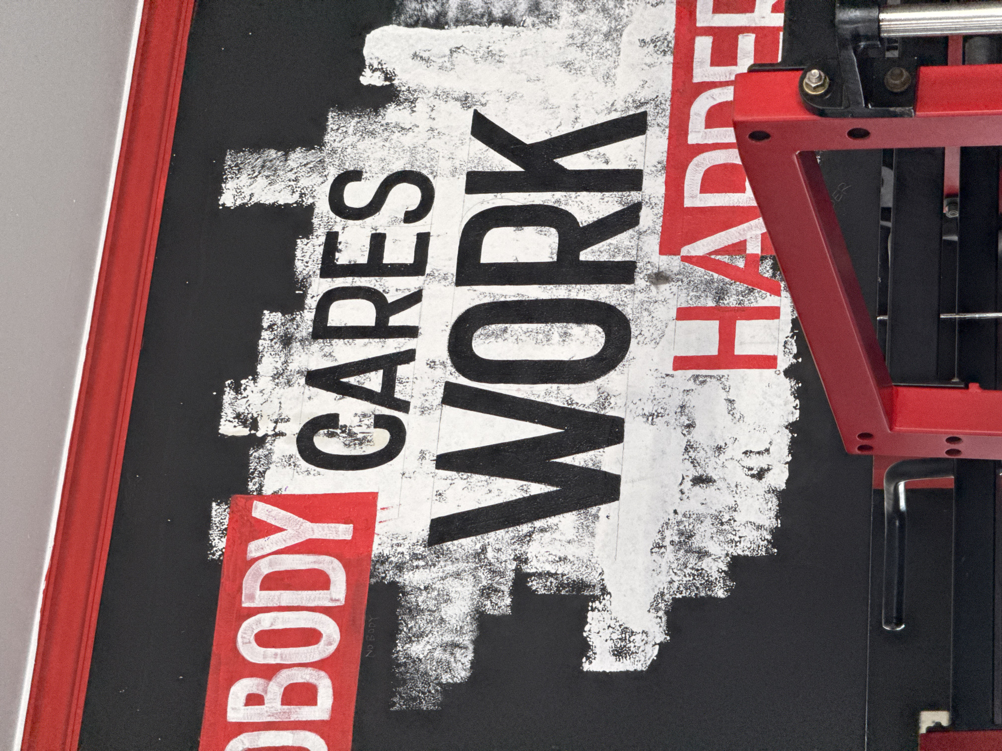 A gym interior in Kuta Selatan, Indonesia, features a motivational mural on a black wall. The mural reads "NOBODY CARES WORK HARDER" in large, bold letters. "NOBODY" is displayed in white text on a red rectangular background in the top left corner. The words "CARES" and "WORK" are in black, with "WORK" being significantly larger and centered. "HARDER" is in white text on a red rectangular background at the bottom right. The mural has a distressed, textured appearance with white brush strokes. Partially visible in the foreground is red gym equipment, possibly a weight rack. The lighting is bright, suggesting an indoor setting.