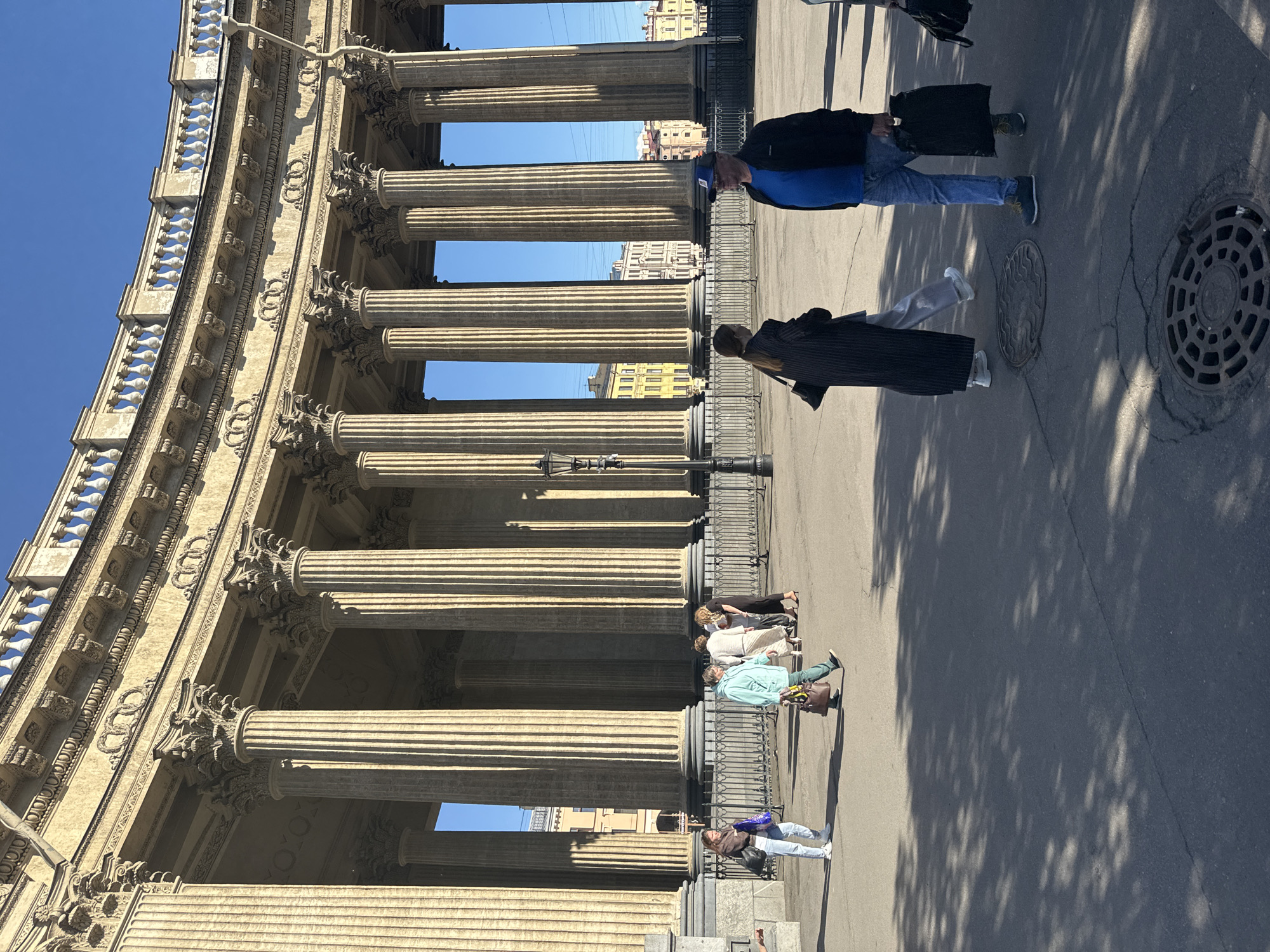 The image shows the Kazan Cathedral in Saint Petersburg, Russia. A group of people are walking in front of the grand colonnade of the cathedral. The architecture is classical, with tall Corinthian columns and ornate carvings. The sky is clear and blue, and the sun is casting shadows on the pavement.
On the left, a woman in a brown jacket and blue jeans walks with a blue shopping bag. Further in the background, a group of people are gathered, some walking and some talking. A woman in a light blue jacket and green pants is carrying a large brown bag.
In the foreground, a man wearing a blue t-shirt, blue jeans, a dark jacket, and a baseball cap is walking towards the right, carrying a black bag. To his left, a woman in a long black coat and light-colored pants is walking away from the camera. The ground is paved with asphalt and has several manhole covers. The overall atmosphere is bright and sunny, suggesting a pleasant day for sightseeing.