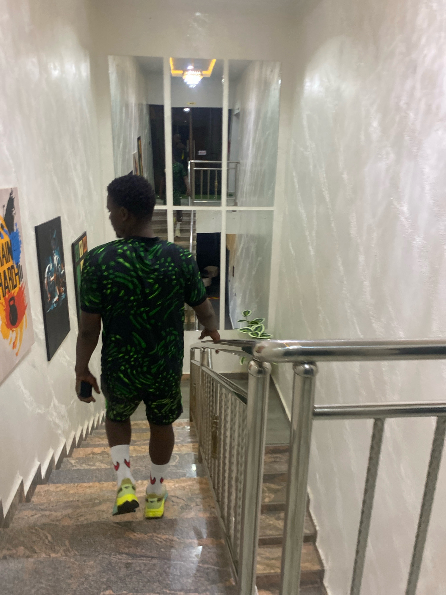 A Black man, seen from behind, walks down a marble staircase in what appears to be a residential building. He is wearing a black t-shirt with a green abstract pattern, matching shorts, white socks with red heart designs, and lime green sneakers. He holds a black smartphone in his left hand. The walls of the staircase are textured and white, with a metallic railing on the right side. The staircase appears to be an interior hallway. The scene does not provide any identifiable markers to place it within Barden Yana, Nigeria.