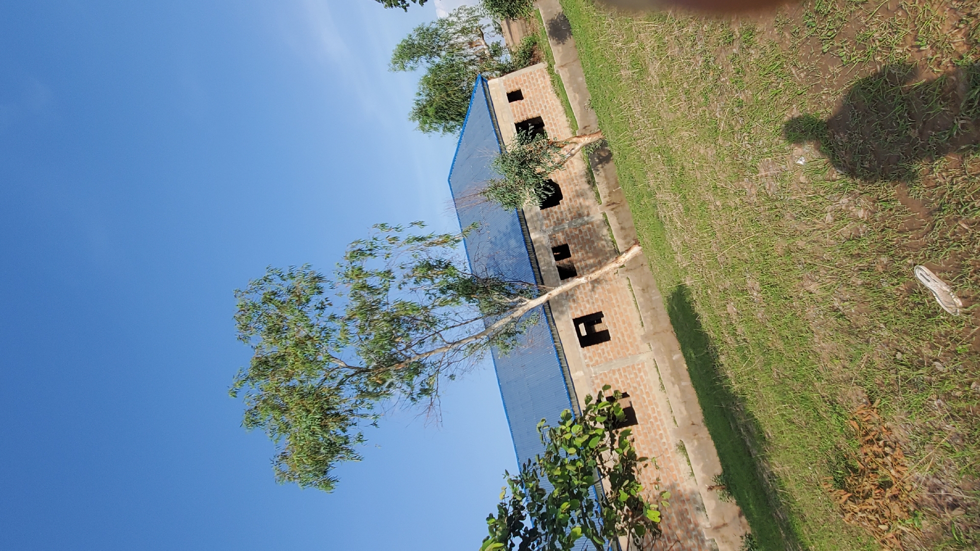 The image depicts an under-construction building with a blue corrugated metal roof, situated in a rural or semi-rural area with dry grass and scattered trees. The building is constructed with light-colored bricks and has several open window frames. The sky is clear and blue. The scene appears to be in Jalingo, Nigeria, as indicated by the presence of indigenous trees and vegetation characteristic of the region. There are no people visible in the frame, but a shadow of a person is cast on the ground, suggesting the photographer's presence. A single shoe is lying on the ground in the foreground. The image is taken at an angle, giving a slightly tilted perspective of the scene.