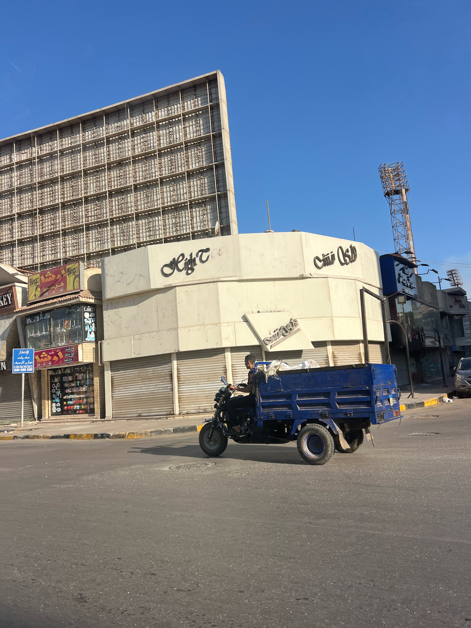 In Giza, Egypt, a blue motorized tricycle with a trailer is driving down a street. The tricycle has a person on it who appears to be a male wearing a black shirt. The street is paved and has yellow and white markings along the side. In the background, there is a large building with a grid-like structure covering its facade, and another curved building with Arabic text signage. The sky is clear and blue. Several storefronts with rolling metal shutters are visible on the ground floor of the buildings. A tall metal tower, likely a light pole, stands in the distance.