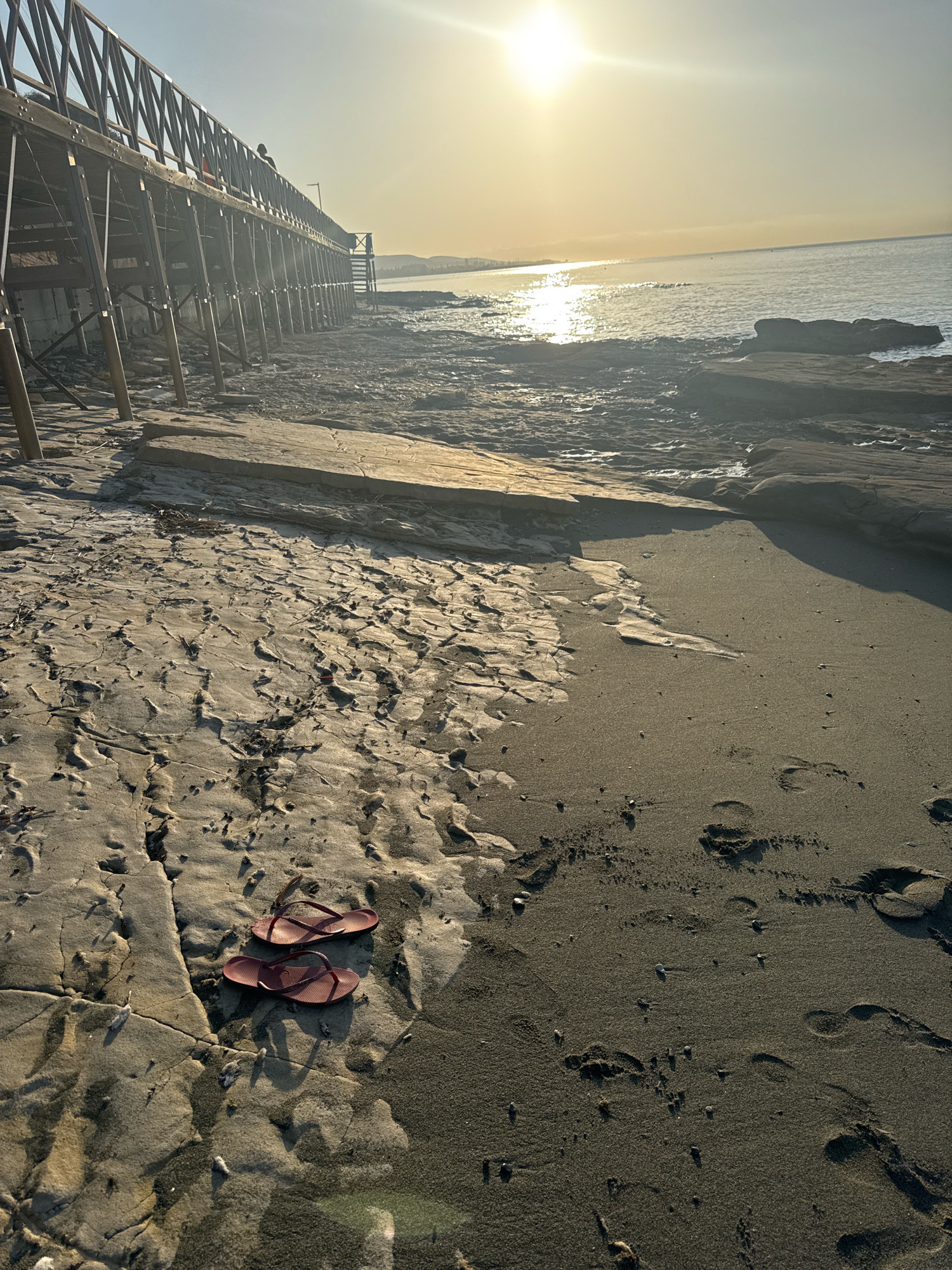 This is a photograph of a beach in Agios Tychona Community, Cyprus, taken during a sunny day at sunset. A wooden pier or walkway stretches across the top left of the frame, its structure casting shadows on the sand below. The foreground is dominated by the beach, with a pair of red flip-flops resting on a rocky, textured patch of sand. Footprints are visible in the darker sand to the right, indicating recent human presence. The sea extends to the horizon, with the sun glinting off its surface, creating a bright reflection. The water appears calm, with some rock formations visible along the shoreline. The sky is clear, with a warm, golden hue from the setting sun.