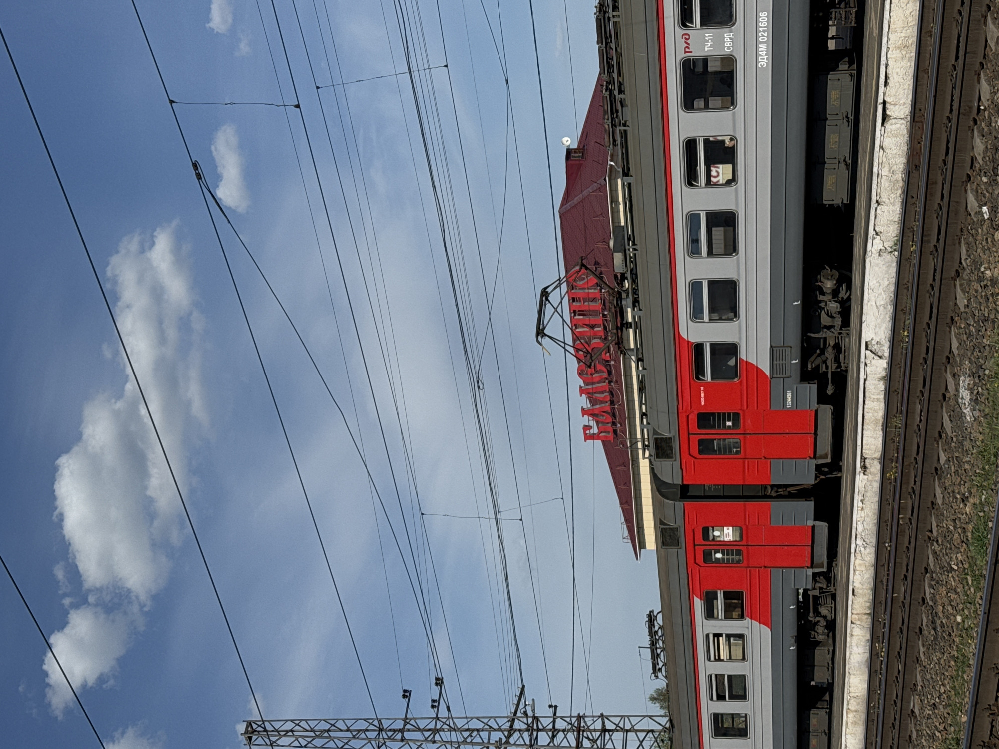 An outdoor shot of a red and grey passenger train with the Russian Railways (RZD) logo, stationary at a station in Balezino, Russia. The train's pantograph is visible, connected to overhead power lines against a cloudy blue sky. The number "3D4M 021606" is printed on the train car. Train tracks run along the platform edge.