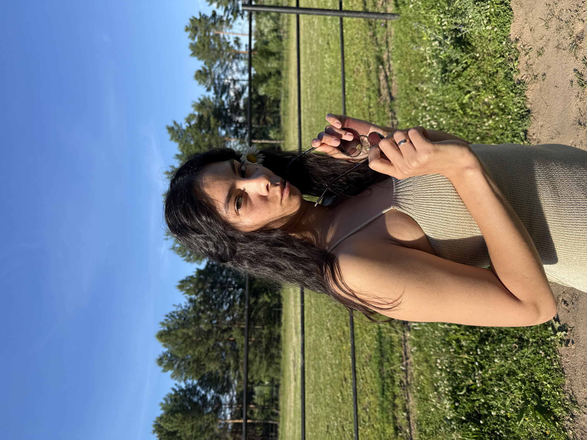 A young woman with long, dark, wavy hair stands in front of a grassy field with a black fence and trees in the background under a clear blue sky. She is wearing a light-colored, ribbed tank top and holds a pair of sunglasses in her hands. A white daisy is tucked behind her ear. She is looking directly at the camera with a neutral expression. The scene appears to be an outdoor location on a sunny day, possibly in the city of Chaikovsky, Russia, given the trees and rural landscape.