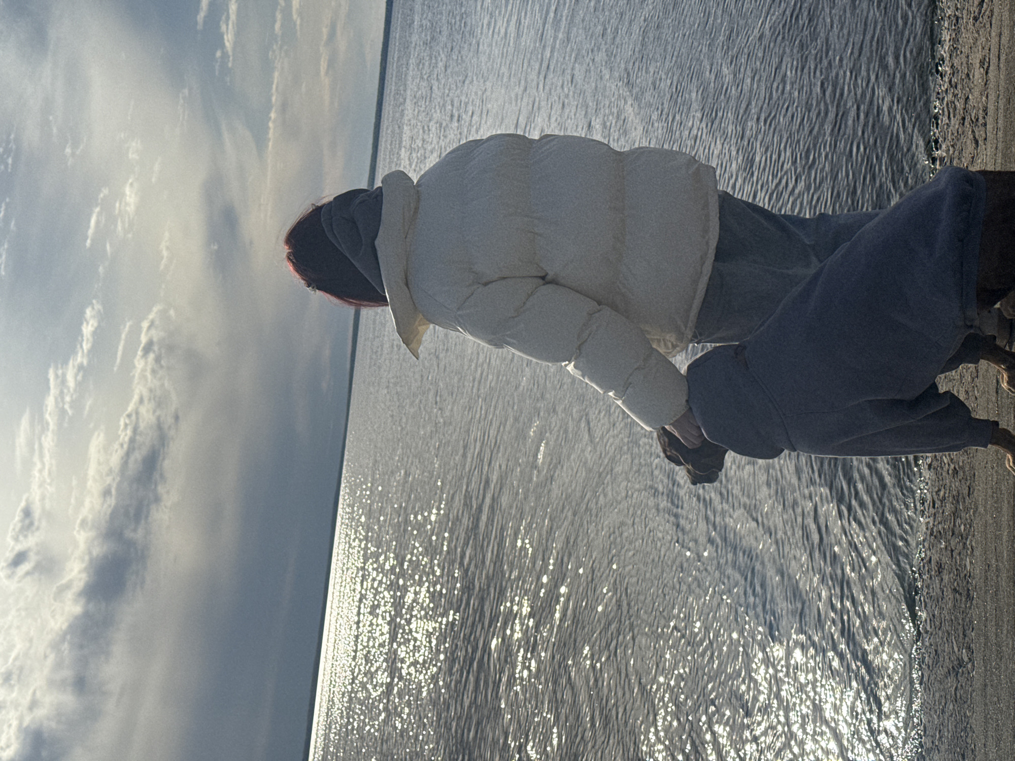 A person in a white puffer jacket and grey sweatpants stands at the edge of a body of water, possibly a lake or river, at Zavolzhye, Russia. The sky is visible with sunlight and clouds. The water surface reflects sunlight, and the person is either touching the water or standing very close to it. The edge of the water meets a sandy or pebbled shore.