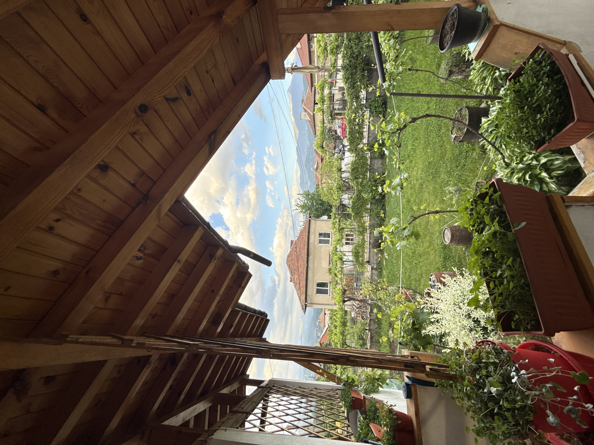 View from a partially enclosed wooden structure, likely a porch or gazebo, in Polena, Bulgaria, overlooking a landscaped garden and residential area.
**Environment:**
* The sky is visible through the structure's opening, showing blue sky with scattered clouds.
* A lush garden is visible with green grass, various plants, and trees.
* A few buildings with red-tiled roofs are scattered in the background, indicating a residential area.
* Mountains are visible in the far distance, adding depth to the scene.
**Objects:**
* The wooden structure consists of a roof and supporting beams, creating a framed view.
* Potted plants and flower boxes are placed on the balcony.
* Vines and other greenery grow along the structure, partially obscuring the view.
* Garden features like a small wooden barrel and metal poles are visible in the garden.
**Location:**
* Polena, Bulgaria is indicated by the residential architectural style and landscape.
* The scene appears to be taken from a residential property, looking out towards the neighborhood and surrounding nature.