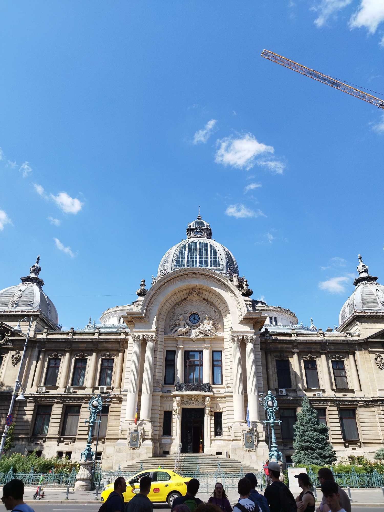 The image shows the National Bank of Romania building in Bucharest, Romania. The building features an imposing classical facade with multiple domes, columns, and ornate architectural details. In the foreground, a yellow taxi cab is parked on the street. Several people are visible on the sidewalk, some looking towards the building, others walking. A construction crane is visible in the bright blue sky. The weather appears to be sunny with scattered clouds.