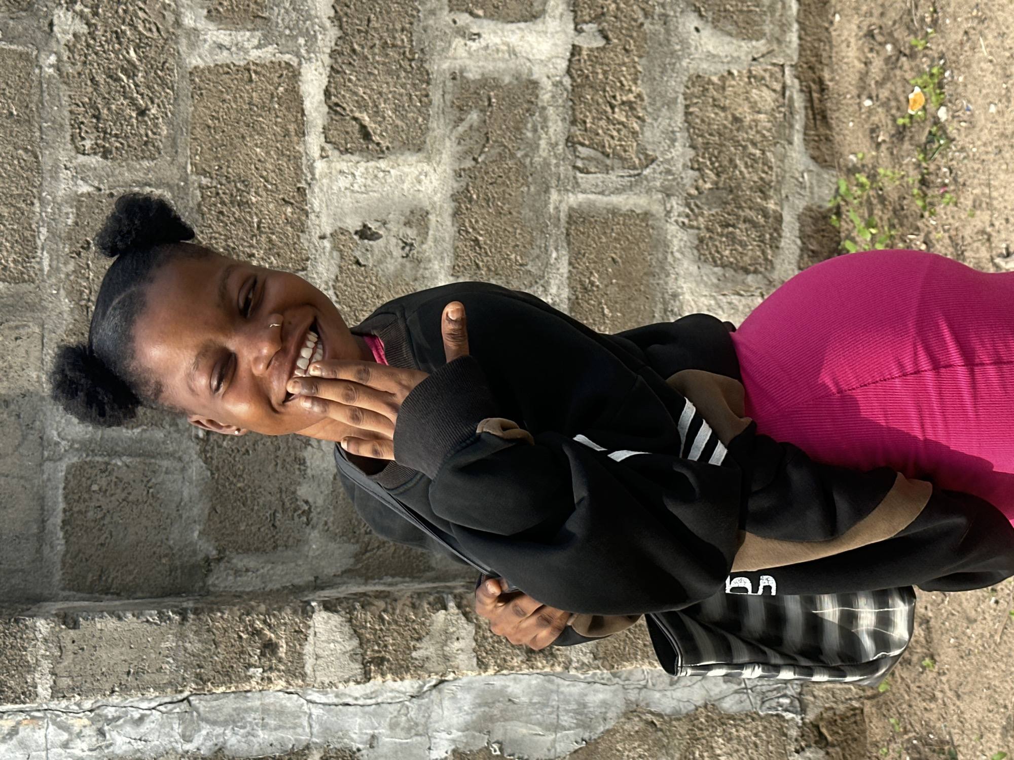 A young Black female with her hair in two buns smiles and covers her mouth with her left hand, her right hand is on her chest. She wears a black hoodie over a pink top and a pink ribbed skirt. A bag with a black and white striped pattern hangs over her left shoulder. She stands in front of a grey brick wall. The ground is dirt with some scattered debris and small green plants. The scene is outdoors in what appears to be an urban or semi-urban environment in Badagry, Nigeria.