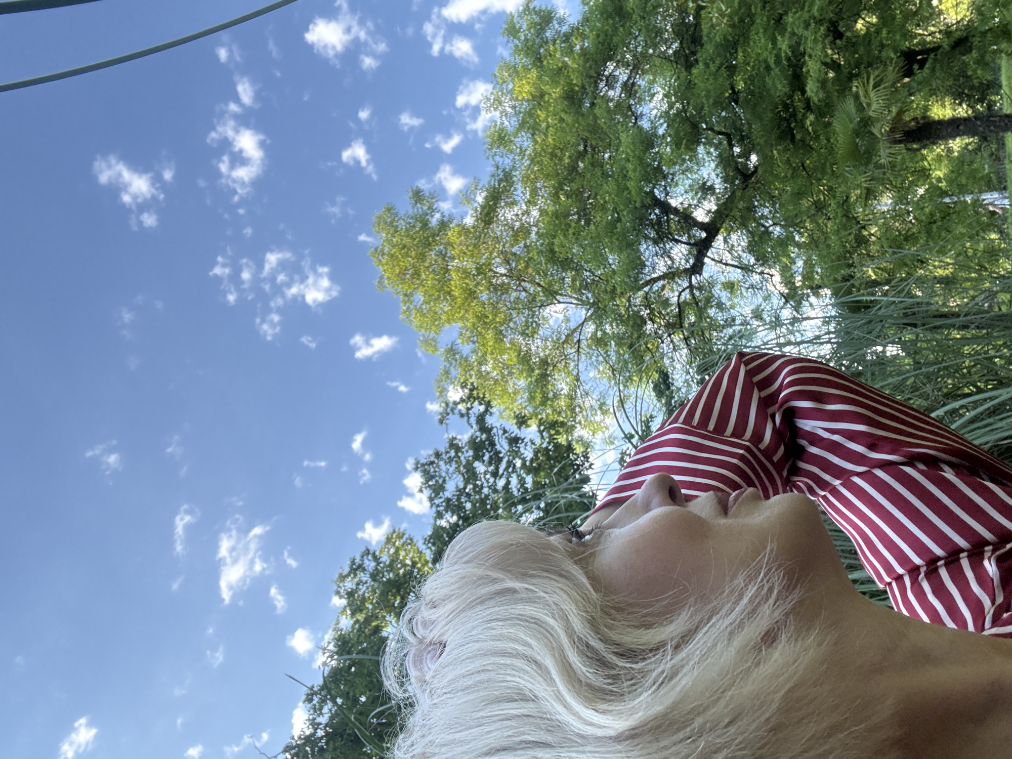 The image shows a close-up profile of a woman with blonde hair, looking upwards. She is wearing a red and white striped shirt. The background consists of lush green trees and a bright blue sky with scattered white clouds. There are no identifiable landmarks of Sochi, Russia in the image.