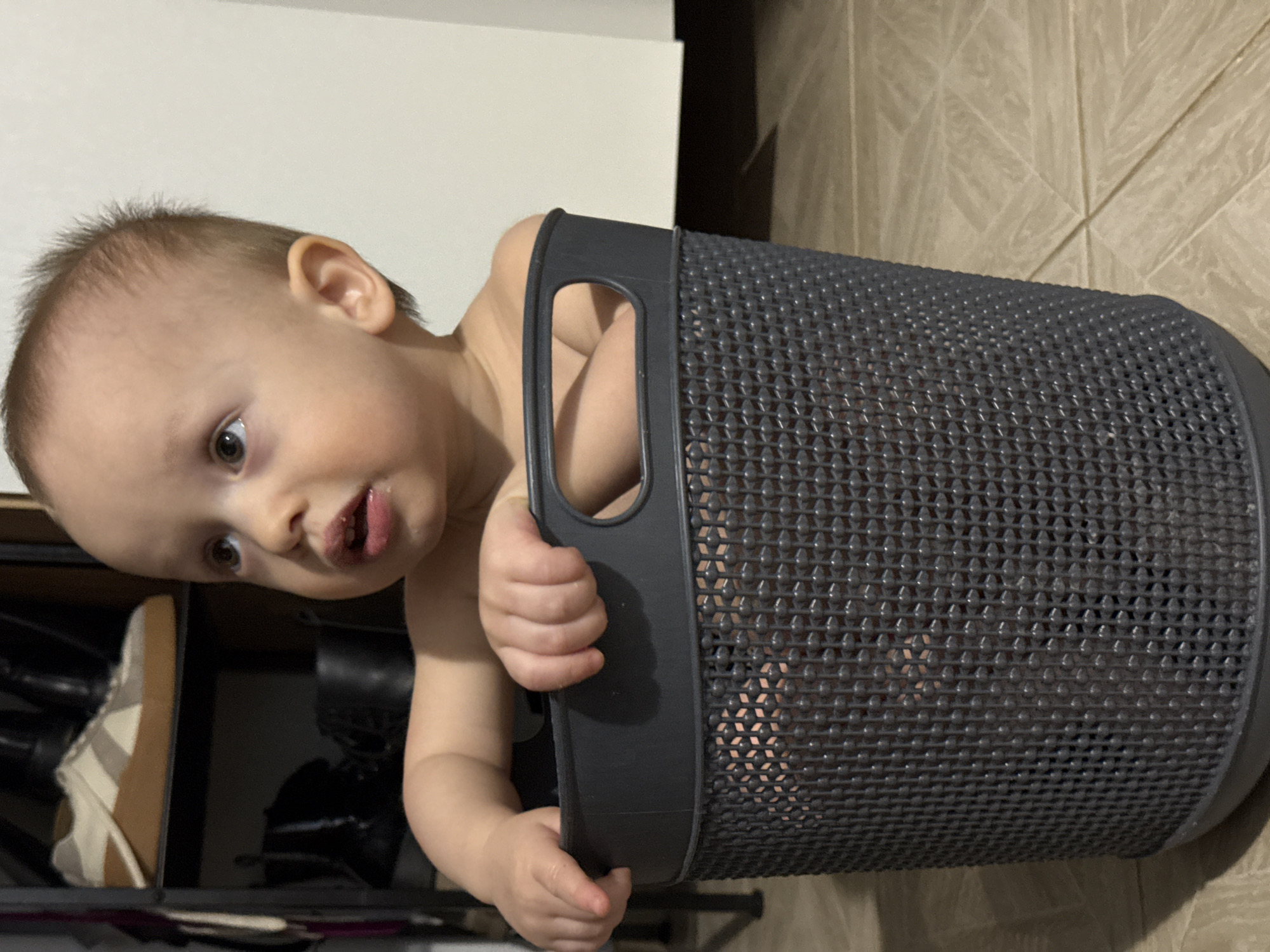 A toddler is partially inside a gray plastic laundry basket. The child is fair-skinned with brown hair. The child's face is tilted towards the camera. The child has brown eyes. The surrounding environment appears to be inside a home. There is tiled flooring. The laundry basket is next to a shelf with shoes.
The picture was taken in Chaikovsky, Russia.