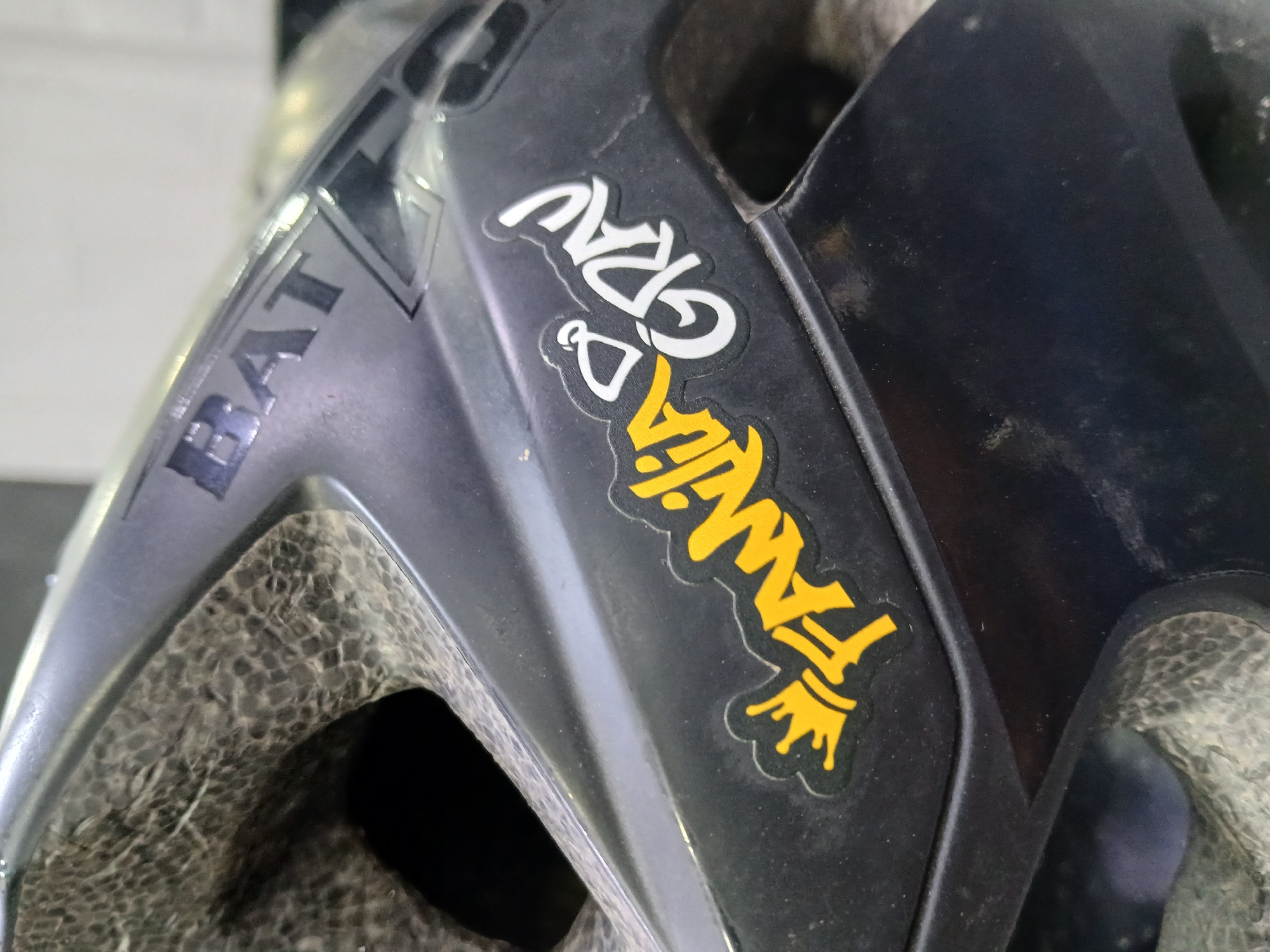 This image shows a close-up of a portion of a helmet. The helmet has the word "BAT" printed on the side in silver. Additionally, there is a sticker with the words "Familia & GRAU" in a stylized font, with "Familia" in gold and "GRAU" in white. The environment is ambiguous, and there's no information suggesting it is in Juan Griego, Venezuela.
