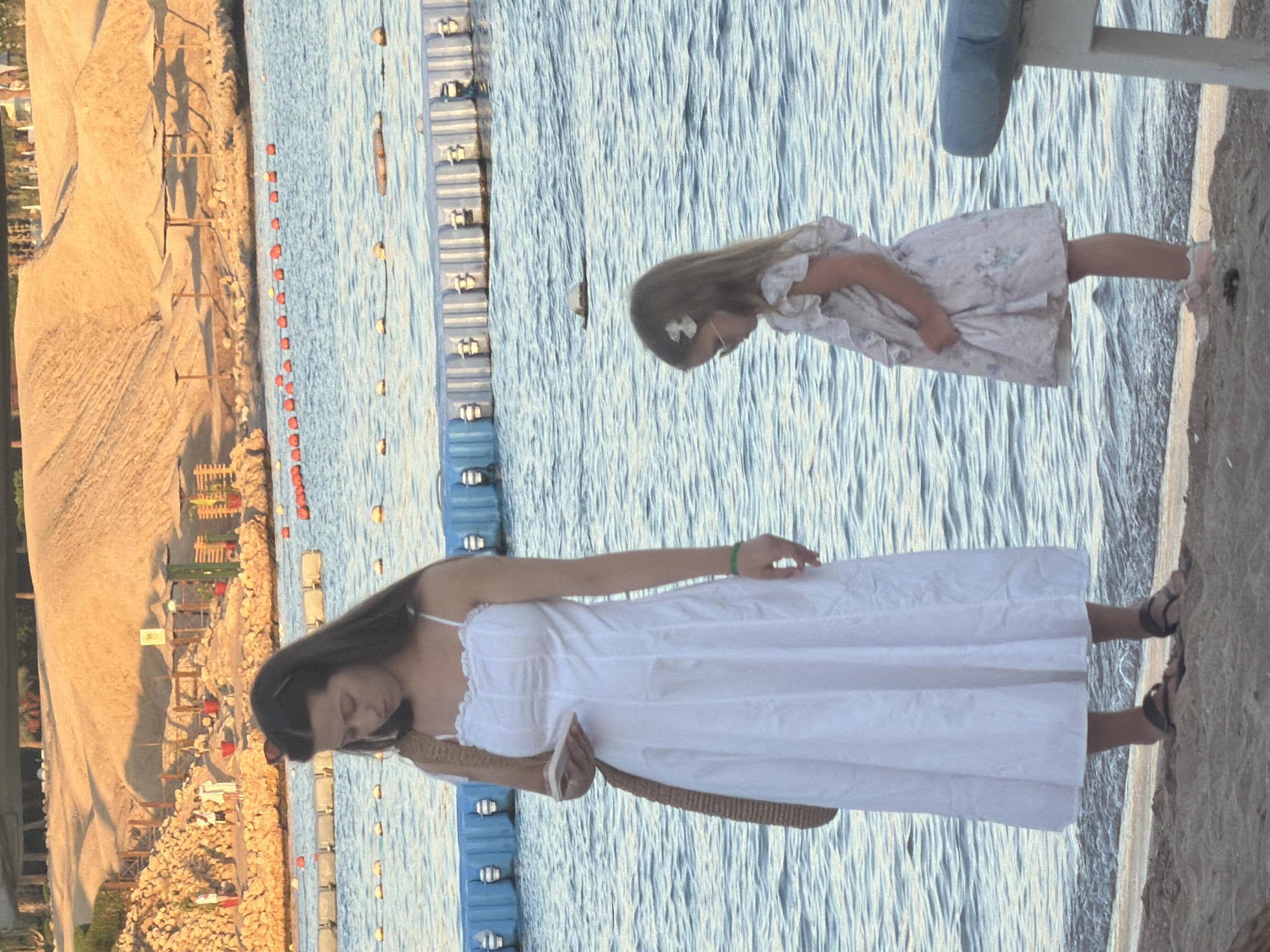 The image depicts a beach scene in Sharm El Sheikh, Egypt, with two females in the foreground. On the left, a young woman with long brown hair, wearing a white sundress and dark sandals, stands on the sand near the water's edge. She is holding a phone in her left hand and has a straw bag slung over her right shoulder. A green bracelet is visible on her right wrist. To her right and slightly in front, a young girl with blonde hair, adorned with a white bow and sunglasses, wears a light-colored floral dress. She stands on the sand as well, with her left hand holding the hem of her dress. In the background, the blue water of the sea is visible, with a row of buoys and floating barriers separating areas of the water. Beyond the water, a sandy hillside is prominent, dotted with sun umbrellas and some vegetation. The overall lighting suggests it is late afternoon or early evening, with a warm glow on the sand.