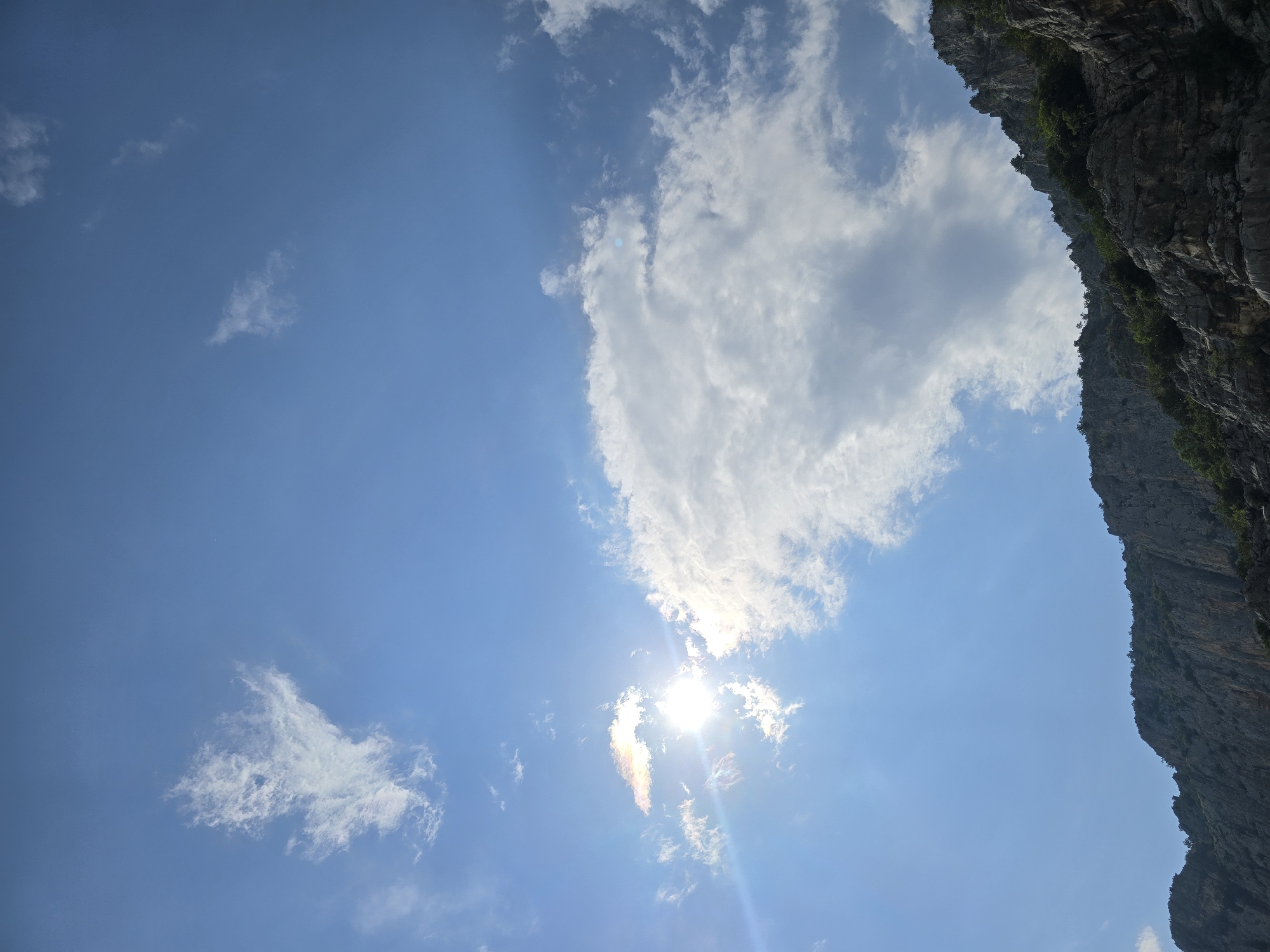 The image shows a dramatic, sun-drenched sky over a rugged, mountainous landscape. The sky is a brilliant blue, dotted with wispy white clouds and larger, puffier formations. The sun, a bright, intense orb, peeks through a cluster of clouds on the left side of the frame, casting a sunbeam that illuminates a rainbow-like halo around it. The lower portion of the image is dominated by the steep, rocky slopes of a mountain range. The rocks are textured and appear weathered, with sparse vegetation clinging to their surfaces. The overall impression is one of natural grandeur and a bright, clear day. The location appears to be a natural, undeveloped area, likely within the mountainous terrain surrounding Podgorica, Montenegro, given the geological features. There are no people or man-made structures visible in the frame.