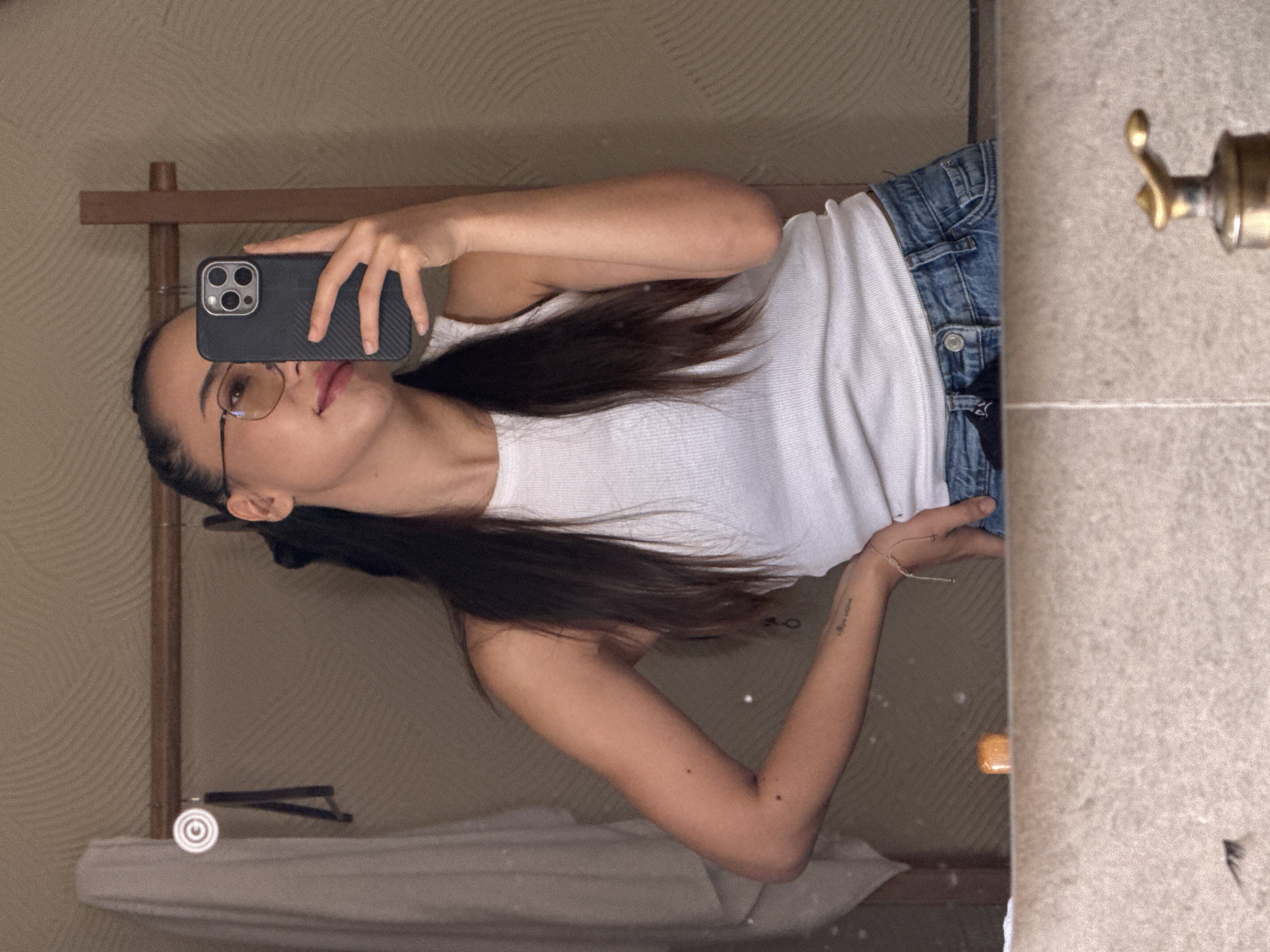 A young woman with dark hair and glasses takes a mirror selfie in a bathroom. She is wearing a white tank top and blue jeans. Her left arm is bent, and her hand is on her hip. A phone is held in her right hand, capturing the reflection. The background of the bathroom features textured walls and a wooden rack with a towel. The image is taken in Da Lat, Vietnam.
