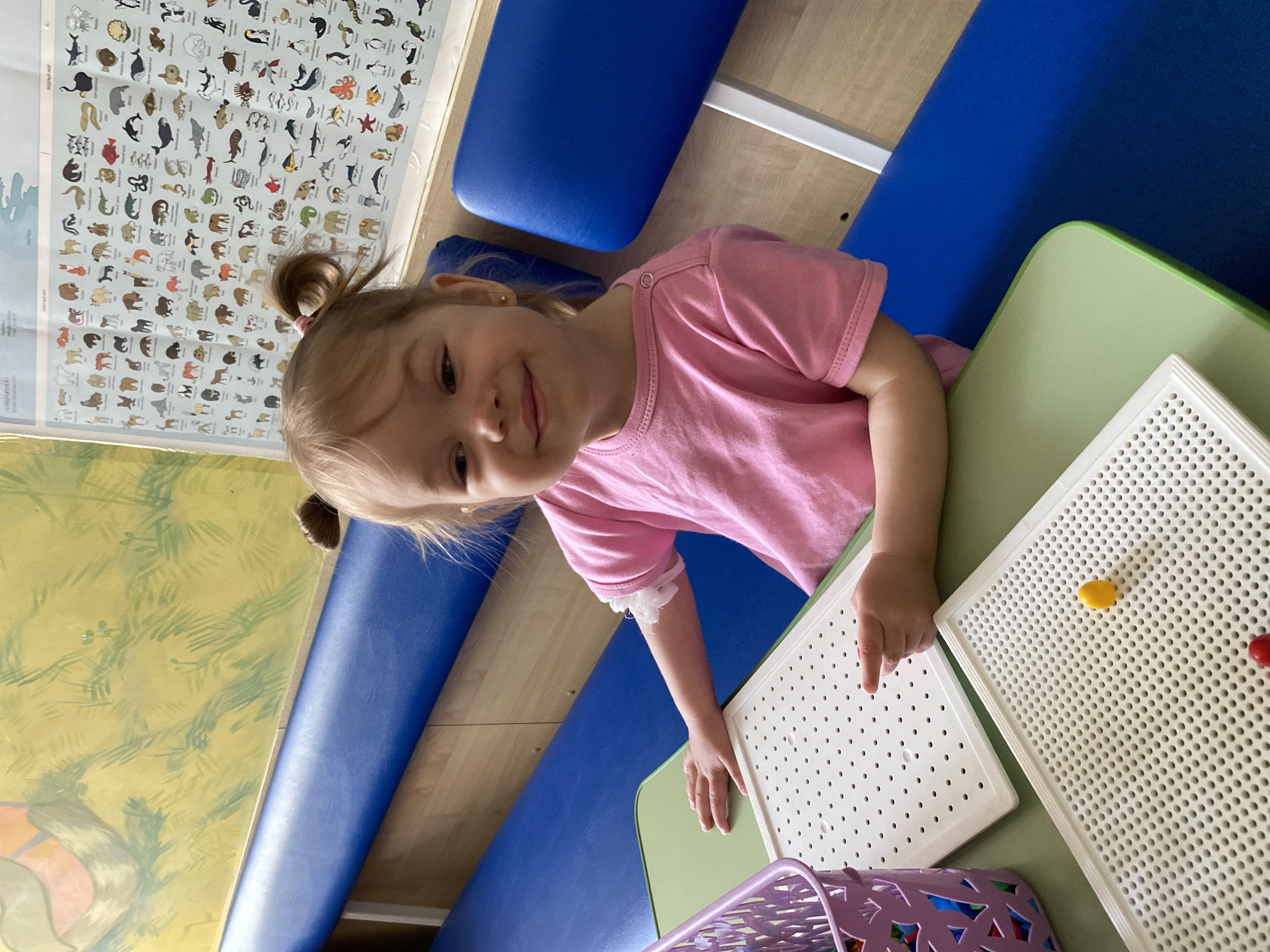 A young girl with blonde hair in pigtails, wearing a pink shirt, is sitting at a green table. She is looking at the camera with a slight smile. On the table are two white plastic boards with small holes, one with a yellow and a red peg in it. A purple basket with colorful objects is at the bottom of the frame. A blue padded bench runs along the back wall. A poster showing various animals is affixed to the wall above the bench. The location is suggested to be in Perm, Russia.