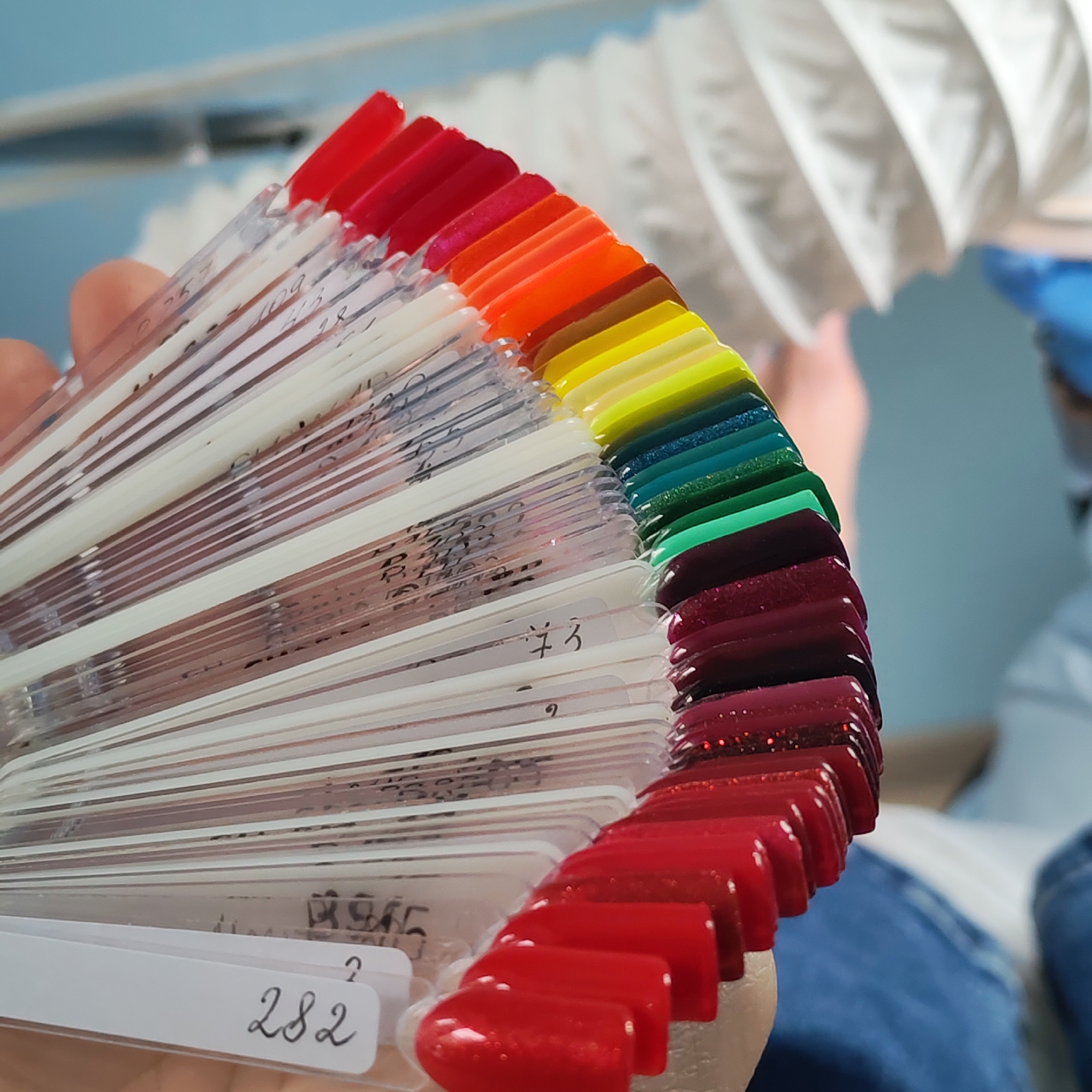 A close-up, low-angle shot features a person's hand holding a fan of nail polish color samples. The samples fan out from the hand, showcasing a gradient of colors from red at the top to a deep green at the bottom, with shades of orange, yellow, and other colors in between. Each sample has a small white label at its base. The background is blurred, showing a white, accordion-like structure and a light blue surface. The image appears to be taken indoors. There is no information in the image to suggest a location in Chaikovsky, Russia.