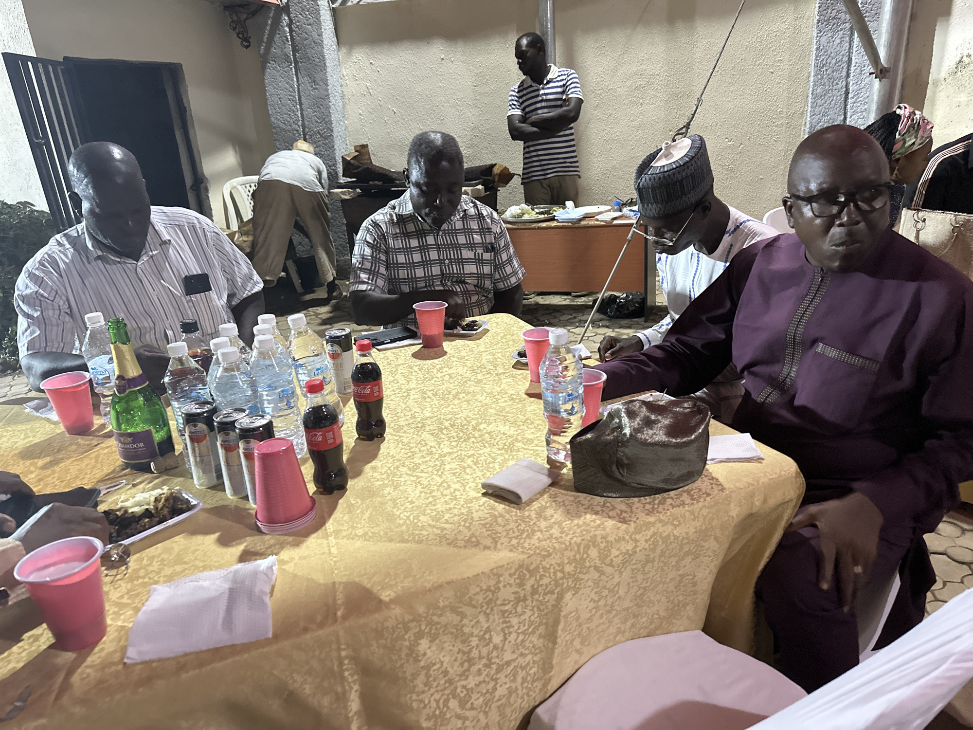 In Narayi, Nigeria, a group of men are gathered around a table covered with a yellow patterned tablecloth. The table is laden with beverages, including water bottles, Coca-Cola, cans of Amstel beer, and a bottle of green Lamdor. There are also red plastic cups, a plate of food, and napkins. The men are dressed in various outfits, including patterned shirts, a striped shirt, and a purple robe. Some are seated at the table, while others are standing or moving around in the background. The scene appears to be an informal gathering, possibly a meal or social event. The environment is a partially covered outdoor space with a wall in the background.