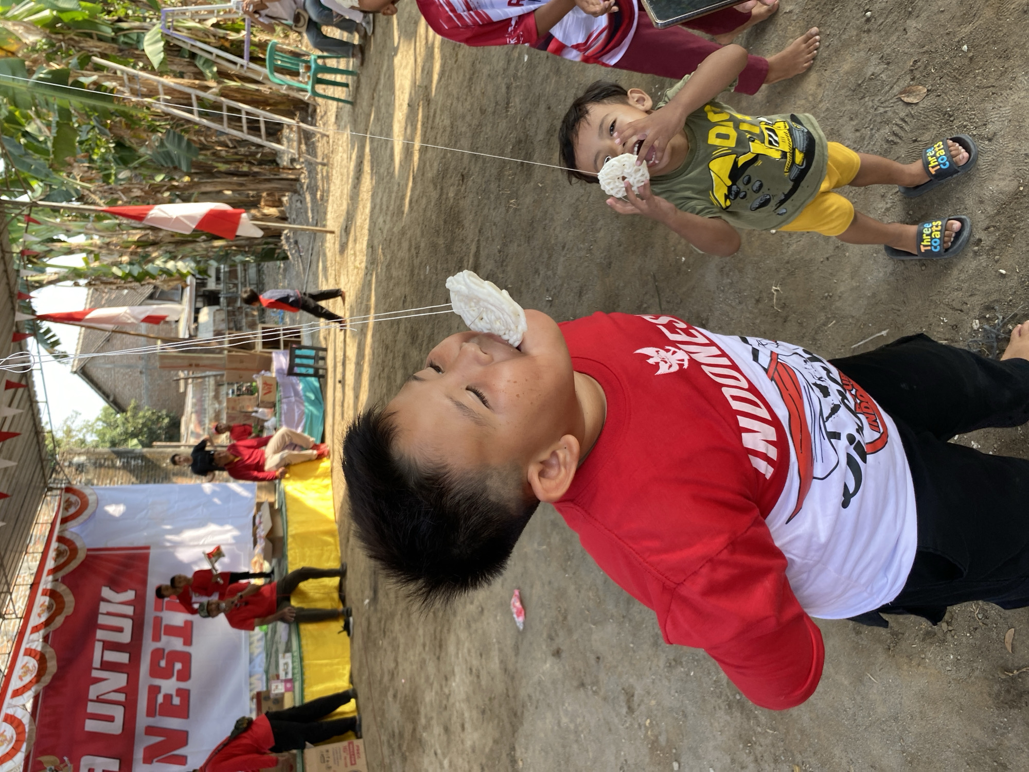 This is a medium shot, eye-level, outdoor photograph capturing a celebratory event, possibly a local festival, in Wonosari, Indonesia. The foreground features two young boys participating in a game involving eating crackers suspended by string. The boy on the left, wearing a red and white t-shirt with "INDONESIA" emblazoned on it, has a cracker in his mouth and his eyes are closed. To his right, a younger boy in a green t-shirt with a yellow construction vehicle graphic and yellow shorts, is holding a cracker up to his mouth. Both boys are barefoot.
In the mid-ground, a stage is set up with a large banner featuring red and white lettering that partially reads "UNTUK INDONESIA". Several adults, mostly wearing red shirts, are present on and around the stage. Some are seated, while others stand. Red and white Indonesian flags are visible in the background, some hanging from poles, and others attached to a ladder.
The environment is sandy and dusty, with some vegetation, including banana trees, visible. The lighting suggests it is daytime, with sunlight casting shadows. The overall atmosphere appears festive and communal.