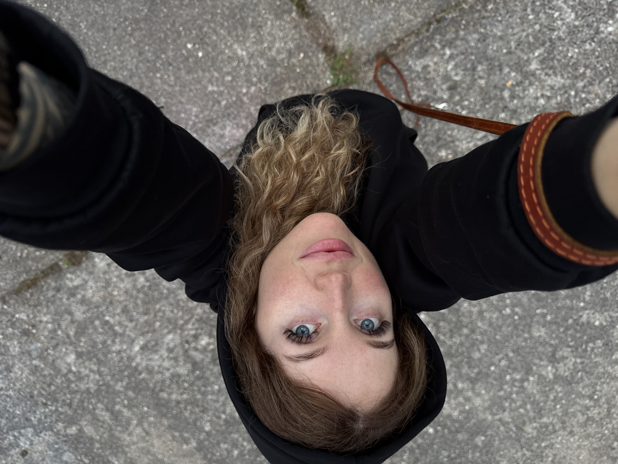 A high-angle selfie shows a young Caucasian woman with blue eyes, blonde wavy hair, and fair skin, looking up at the camera. She is wearing a black hooded sweatshirt. Her expression is neutral. She has noticeable makeup on, including mascara and lip gloss. The background appears to be a grey, textured surface, possibly pavement, with some small patches of green growth visible. A thin, brown strap, possibly from a bag, is visible across her chest and extends downwards. The image was taken outdoors, in daylight, on a sidewalk or public walkway. The specific location is not discernible from the visual elements.