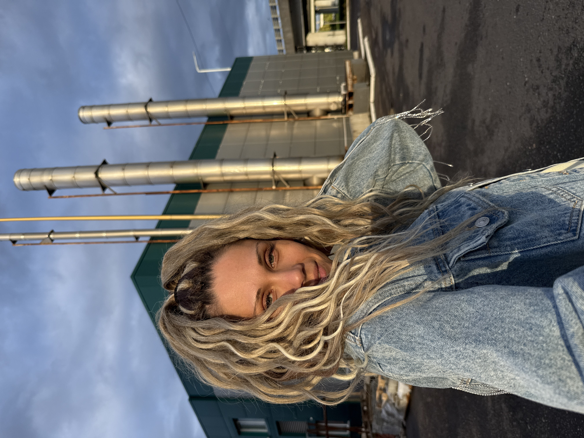 A young woman with blonde, curly hair is standing outdoors in front of a building with industrial smokestacks. She is wearing a denim jacket and is looking at the camera. The sky is cloudy and the sun is setting, casting a warm glow on the scene. The building appears to be an industrial facility, possibly a power plant or factory, with tall metal pipes extending from the roof. The area around the building is paved and appears to be an industrial or commercial zone. The location is described as being in the city of Ignatovo, Russia.