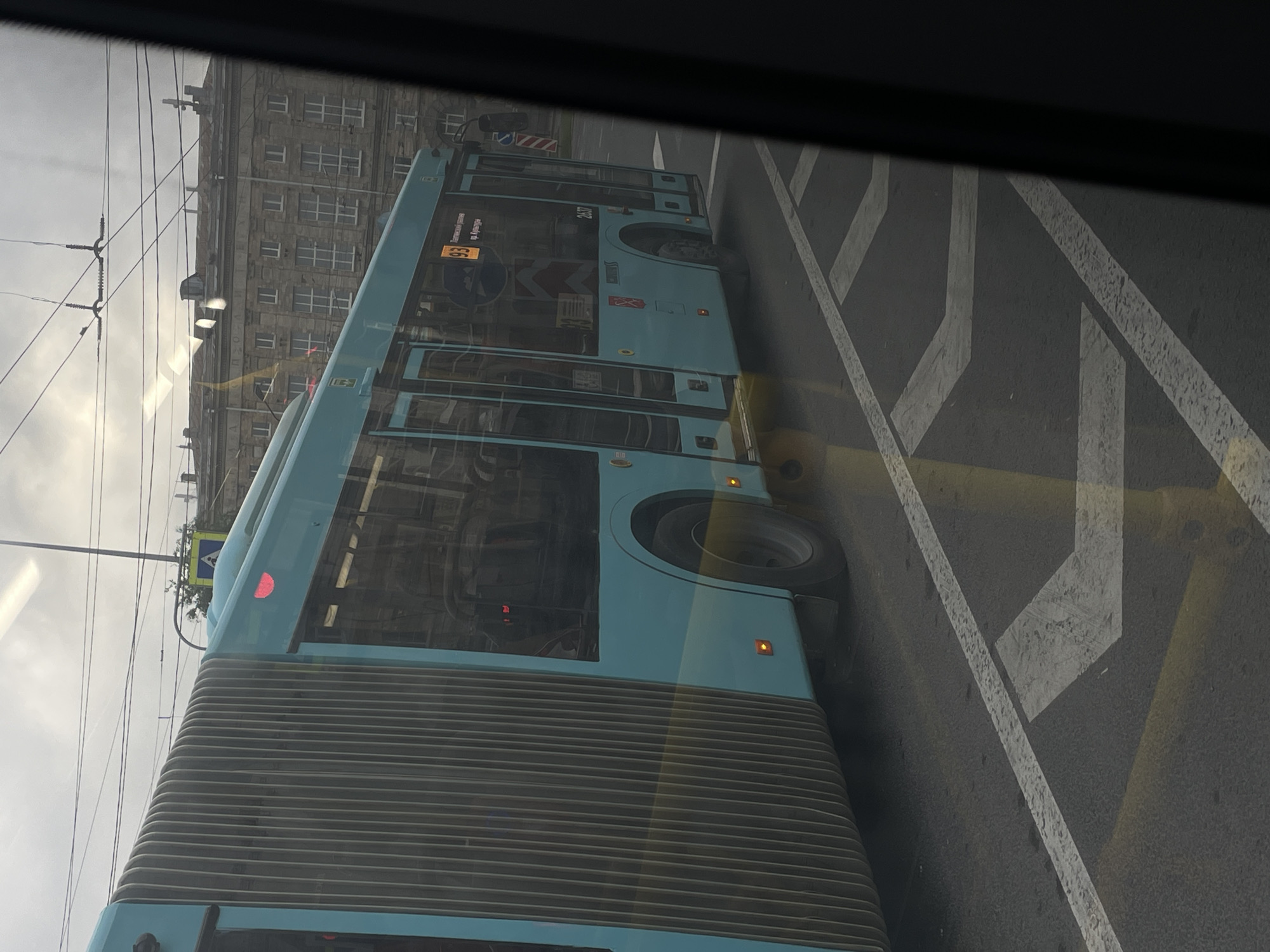The image shows a turquoise articulated bus, identified as route number 93, driving on the street in Saint Petersburg, Russia. The bus is a modern, low-floor model with a large, ribbed bellows section connecting its two parts. It has numerous windows and multiple doors. The route number "93" is clearly visible on the side of the bus in a yellow box. Text beneath the route number appears to be in Cyrillic script, likely indicating the destination or route name. The bus is traveling on a paved road with clear lane markings, including white lines and yellow dashed lines, indicating it is in traffic. In the background, a historic building with multiple windows and a slightly overcast sky are visible. The overall lighting suggests it is daytime, possibly late afternoon or early morning, given the soft light. The perspective is from inside another vehicle, looking out through a window, as indicated by the dark frame in the foreground.