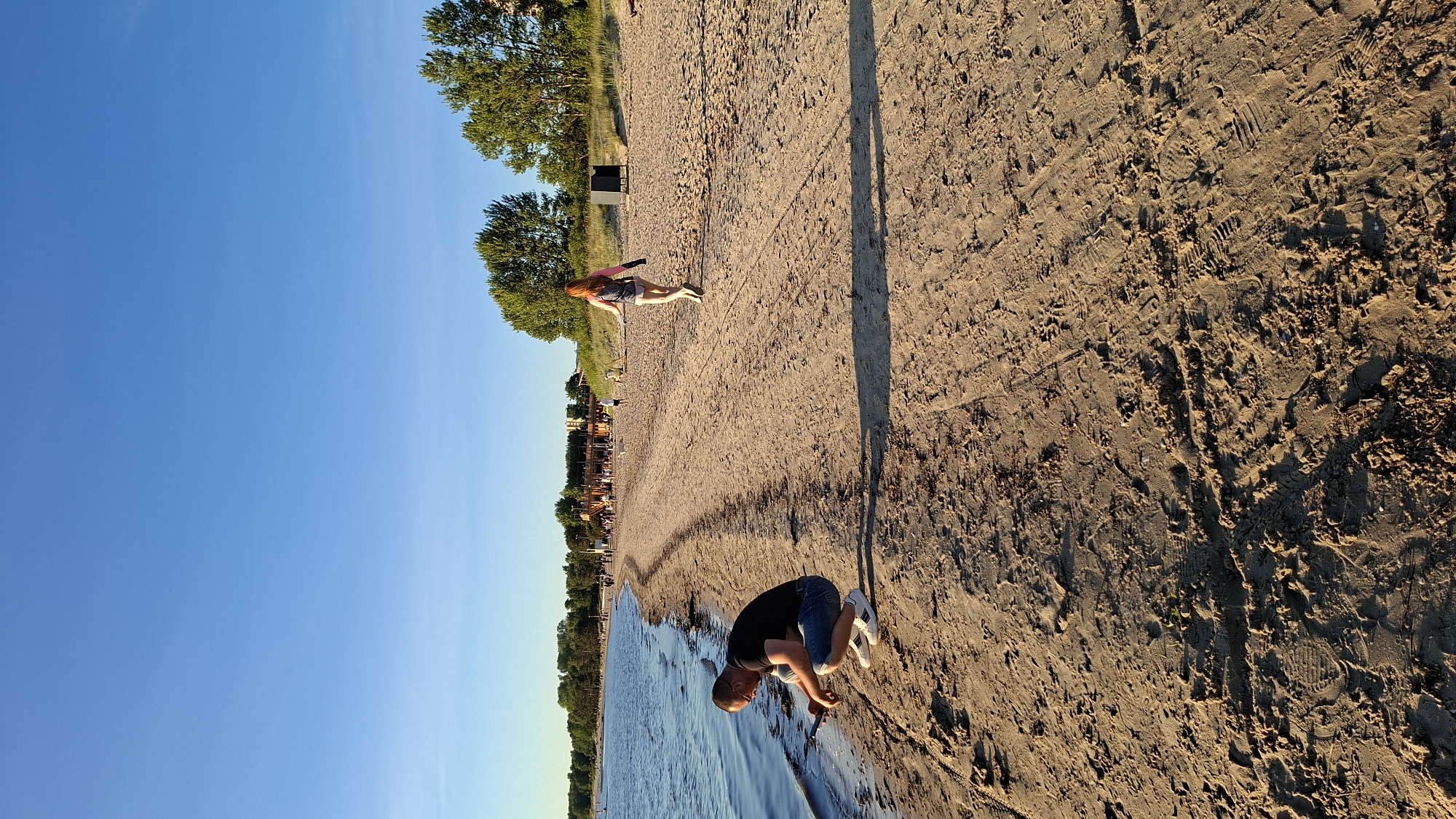 The image depicts a sunny day at Pirita Beach in Tallinn, Estonia. In the foreground, a man wearing a black t-shirt and denim shorts is squatting by the water's edge, looking down at his phone. Further down the sandy beach, a woman with reddish hair, wearing a grey tank top and light-colored shorts, is walking away from the camera, with a backpack on her shoulders. In the background, there are trees, buildings, and other beachgoers, indicating a populated public space. The sky is clear and blue, and the sun casts long shadows on the sand.