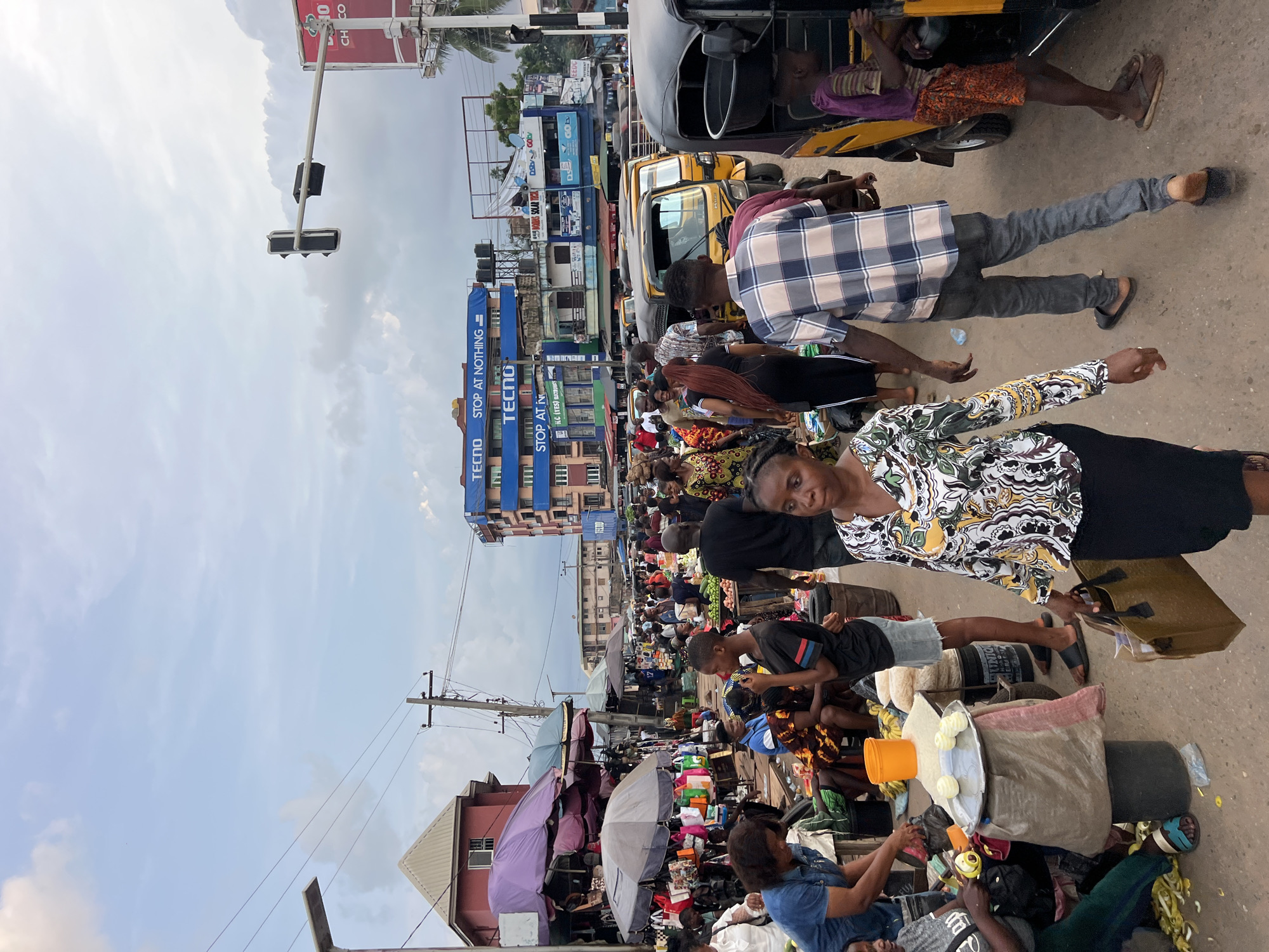 This is a medium shot, eye-level, exterior, daylight photograph taken in Nkpor, Nigeria. The scene depicts a bustling street market with many people and vehicles. In the foreground, a woman in a black skirt and a patterned blouse walks towards the camera, carrying a brown bag. To her left, a person in a blue denim shirt is visible. To her right, a young boy in a purple and striped shirt stands next to a yellow auto-rickshaw. Behind the boy, a man in a plaid shirt and grey pants walks away from the camera.
The market is filled with stalls covered by umbrellas, displaying various goods, including bags and produce. Several yellow auto-rickshaws are parked or moving through the crowded street. In the background, multi-story buildings with signage for "TECNO" are prominent. Traffic lights are visible overhead, indicating a busy intersection. The sky is cloudy with patches of blue. The overall impression is one of a lively and active commercial area.