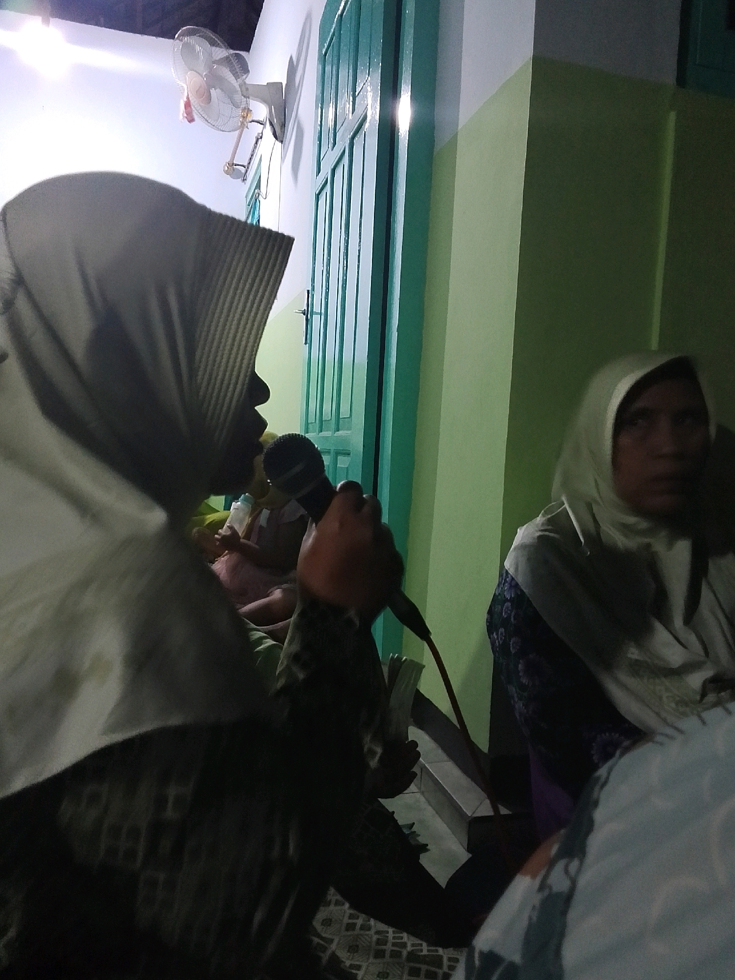 The image depicts an indoor scene, possibly a gathering in Bulurejo, Indonesia. In the foreground, a person wearing a hijab is holding a microphone and speaking. To their right, another person also wearing a hijab is seated. Behind them, a child is visible, possibly being held. The background shows a green door and a wall painted in light green and white. A fan is mounted on the wall in the upper left corner of the image. The lighting appears to be artificial, creating shadows and highlighting certain areas of the scene. The overall atmosphere suggests a casual, possibly familial, event.
