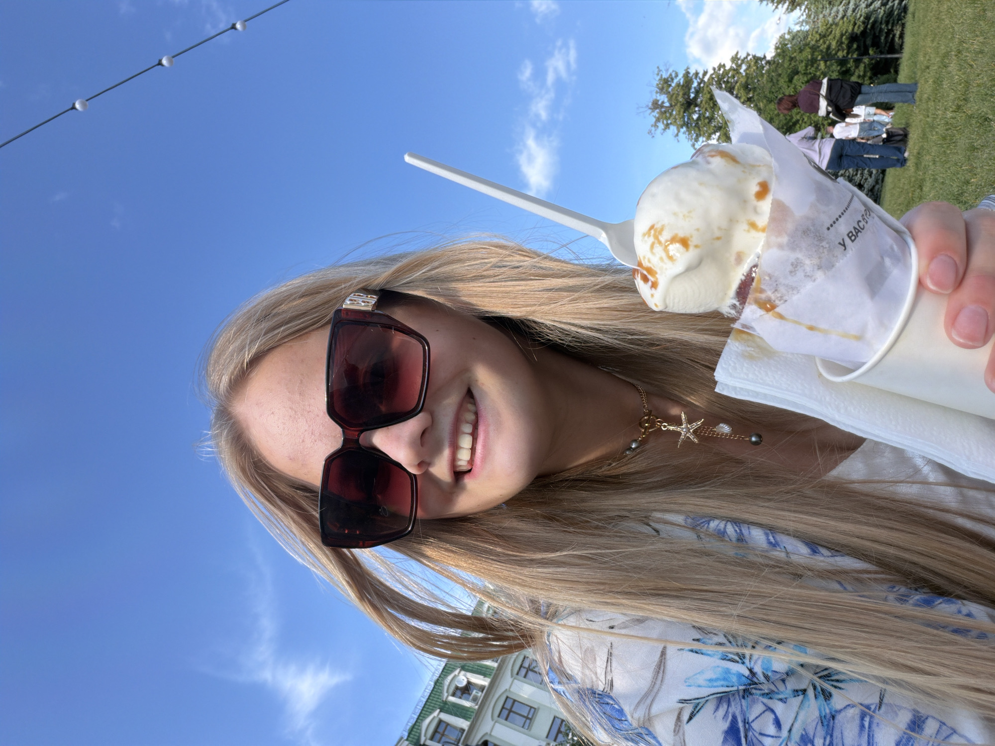 A young woman is depicted in a close-up, front-facing selfie, smiling outdoors under a clear blue sky in Kazan, Russia. She has long blonde hair and wears large dark-rimmed sunglasses. Her attire includes a white top underneath a loose-fitting blue and white patterned blouse or jacket. Around her neck is a gold chain necklace featuring a prominent starfish charm, a smaller shell charm, and two dark pearl beads. She holds a white paper cup in her right hand, containing two scoops of vanilla ice cream generously drizzled with caramel sauce. The ice cream is partially wrapped in a white paper napkin with some indecipherable Russian text, and a white plastic spoon is embedded in it. In the background, to the left, a light-colored building with green roofs and numerous windows is visible. To the right, a grassy park area with various trees and several casually dressed individuals walking or standing in the mid-ground can be seen. String lights are strung across the upper right portion of the sky.