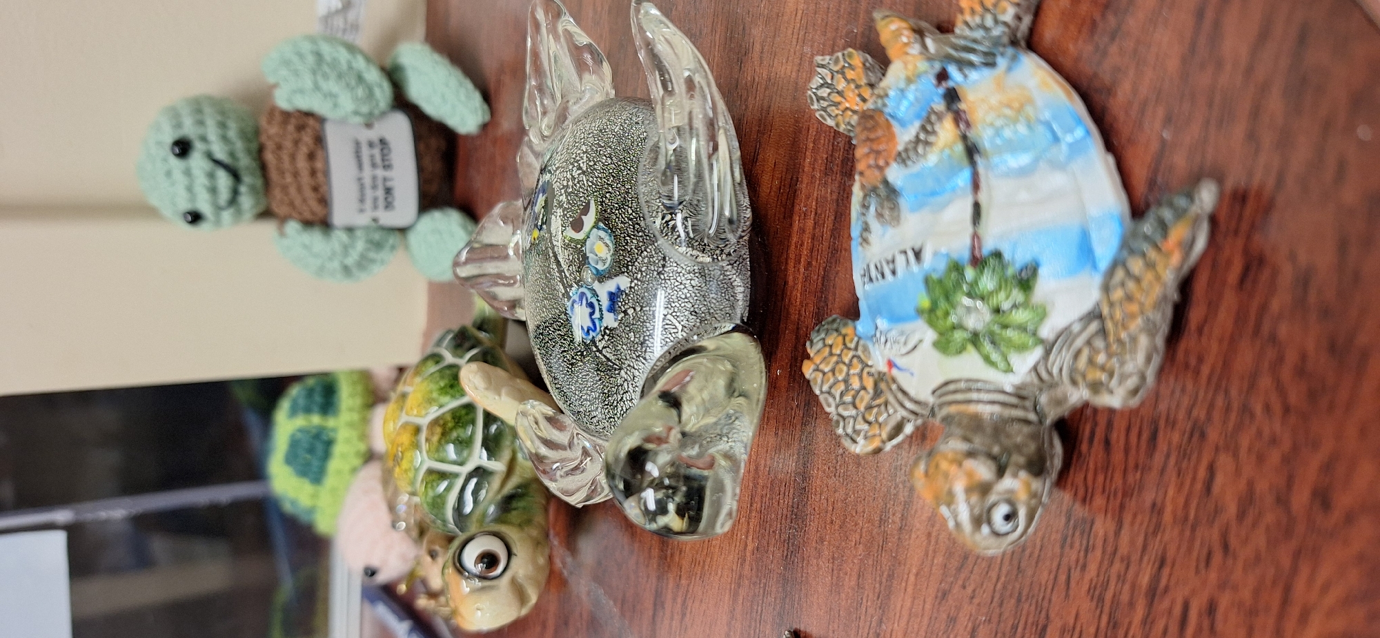 In the city of Upper Tantallon, Canada, a group of turtle figurines are displayed on a wooden surface. The figurines are of varying materials and designs. In the foreground, a detailed figurine of a turtle with a colorful shell depicting palm trees and a tropical landscape rests on the wooden surface. Behind it, a clear glass turtle figurine with intricate colorful designs embedded within the glass is positioned. Further back, a ceramic turtle figurine with large, expressive eyes and a green and yellow shell is visible. In the background, a light green crocheted turtle figure is seen, holding a sign that reads "I don't wanna go but they won't let me DON'T STOP." Another crocheted turtle with a green shell is partially visible to the left, and a pink crocheted object is also present. The lighting in the scene is artificial, casting shadows and highlighting the textures of the figurines. The overall environment appears to be an indoor setting, possibly a desk or shelf.
