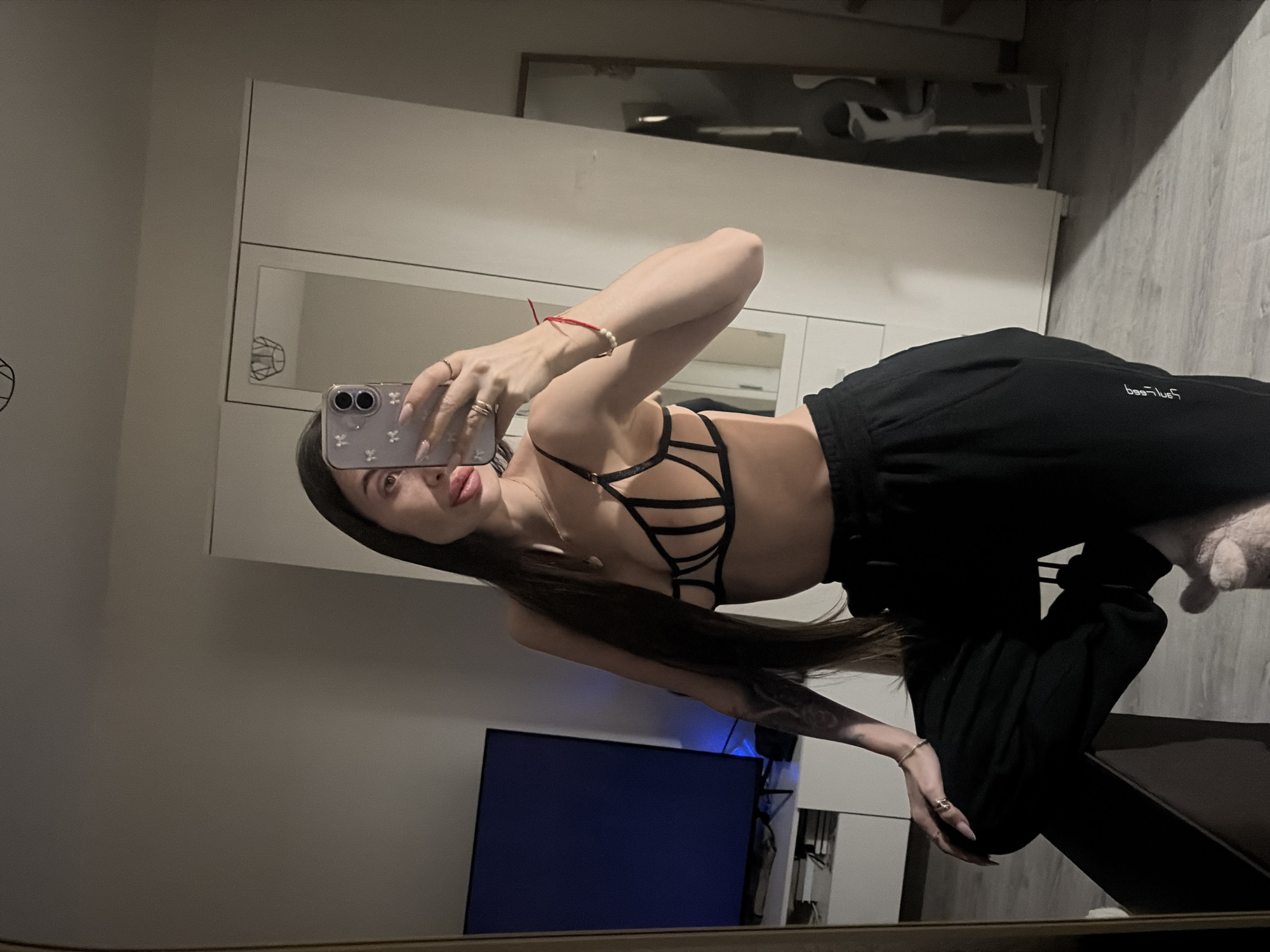 A selfie image shows a woman indoors, presumably in her residence. She is holding a smartphone up to take the photo, partially obscuring her face with the device. She is wearing a black bra-like top with a complex strap design and loose black pants. Her long hair is partially visible.
In the background, there is a television, a white wardrobe, and what appears to be a reflection of another part of the room in a mirror.
Given the context, there is no evidence to suggest the image was taken in Saint Petersburg, Russia.