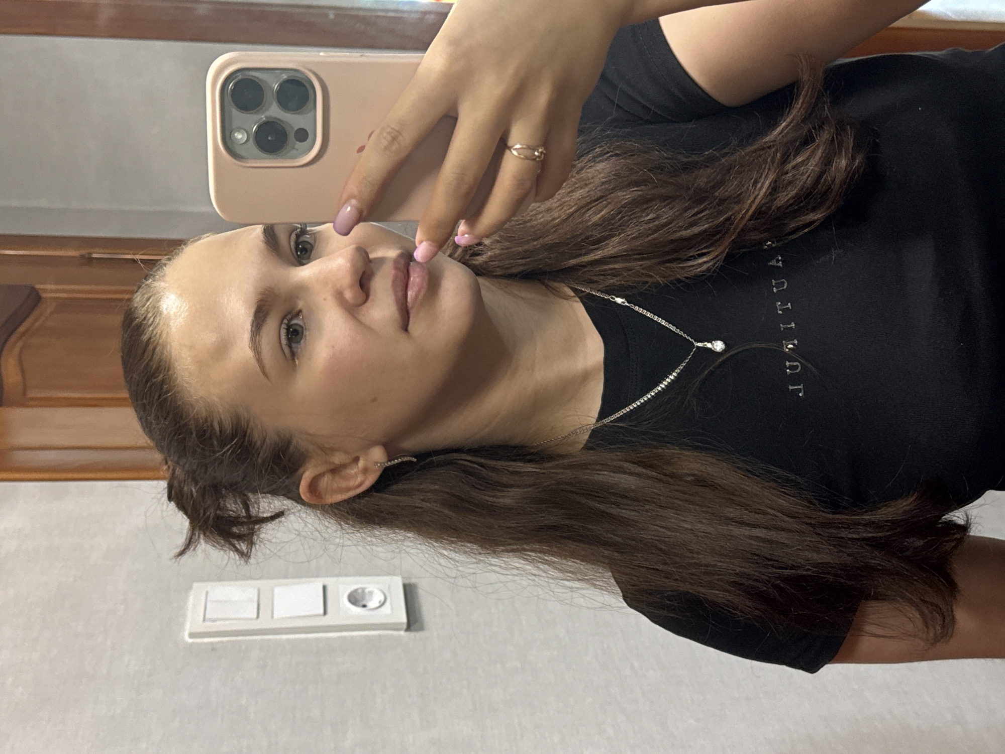 A young woman with light skin and brown hair takes a selfie with a rose-gold iPhone. She wears a black t-shirt and a silver necklace. Her nails are painted a light purple. A light switch and outlet are visible on the wall behind her. The location is likely an indoor setting. There is no specific evidence indicating the scene is in Chaikovsky, Russia.