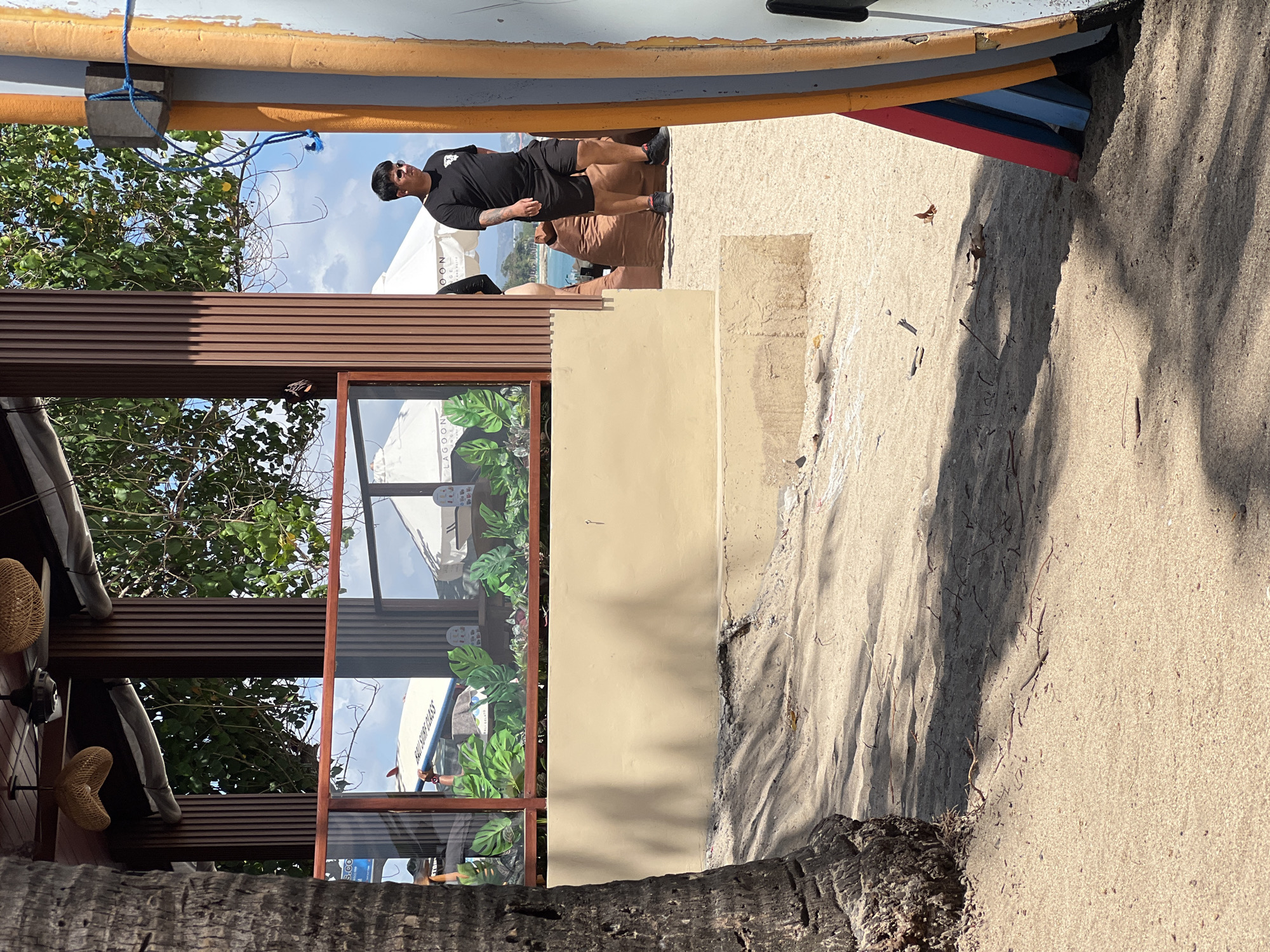 In Kuta, Indonesia, a man in a black t-shirt and shorts walks on a sandy beach near a building with wooden pillars and a glass partition. Green plants are visible behind the glass, and two white umbrellas are set up in the background. The scene includes a palm tree trunk on the left and a colorful surfboard leaning against a wall on the right. The sky is blue with scattered clouds.