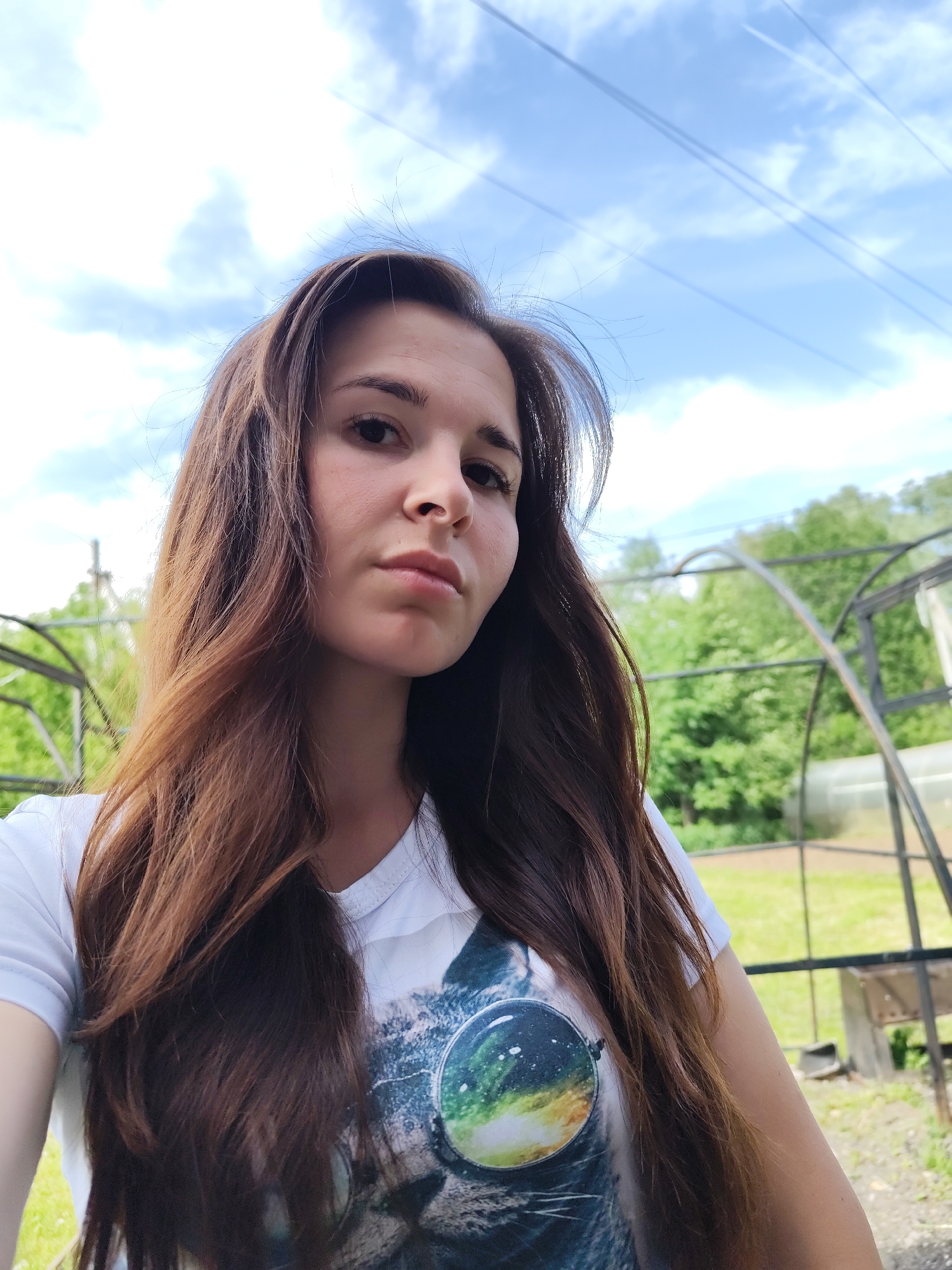 A selfie of a woman with long brown hair, wearing a white t-shirt with a space-themed design. The photo is taken outdoors, with a bright blue sky with scattered clouds in the background. Overhead power lines are visible in the sky. Behind the woman are greenery and a metal greenhouse structure. The location may be in or near Фоки, Russia given the prompt.
