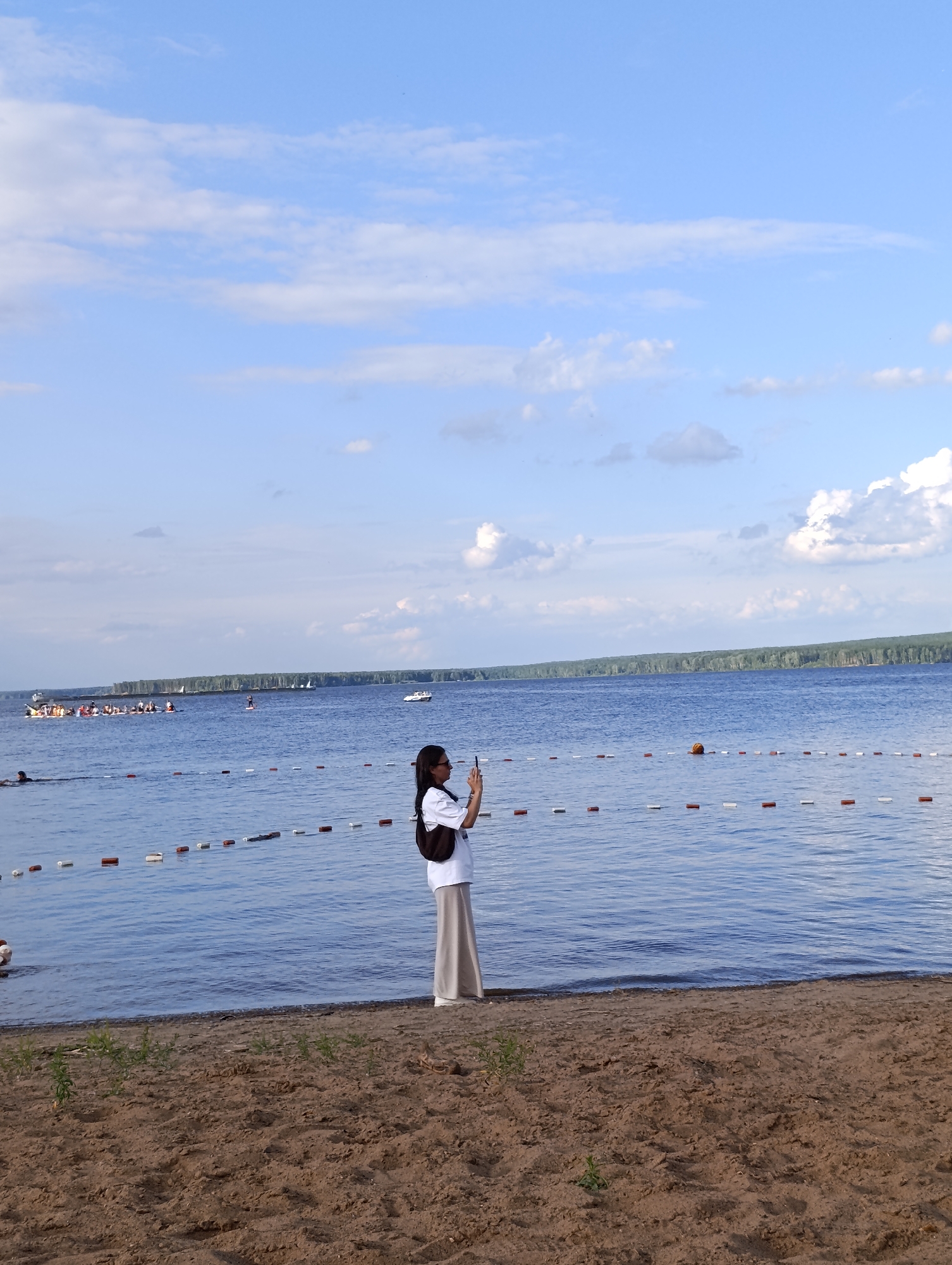 This image shows a serene beach scene with a woman in the foreground taking a photo with her phone. She is dressed in a white shirt and a long, light-colored skirt, and she has dark hair and is wearing glasses. She is standing on a sandy beach, facing the water.
The water is calm and blue, with a line of buoys marking a swimming area. In the distance, there is a long line of trees, suggesting a forest on the opposite shore. Several boats are visible on the water, including a white speedboat and a group of people in kayaks or canoes. The sky is a clear blue with fluffy white clouds, indicating a pleasant day, likely in the afternoon.
The overall mood of the image is peaceful and relaxed, with the woman enjoying the scenery and capturing a moment. The location appears to be a public beach, possibly a resort or a recreational area. The presence of buoys and other boats suggests that this is a popular spot for water activities. There is no visible text in the image.