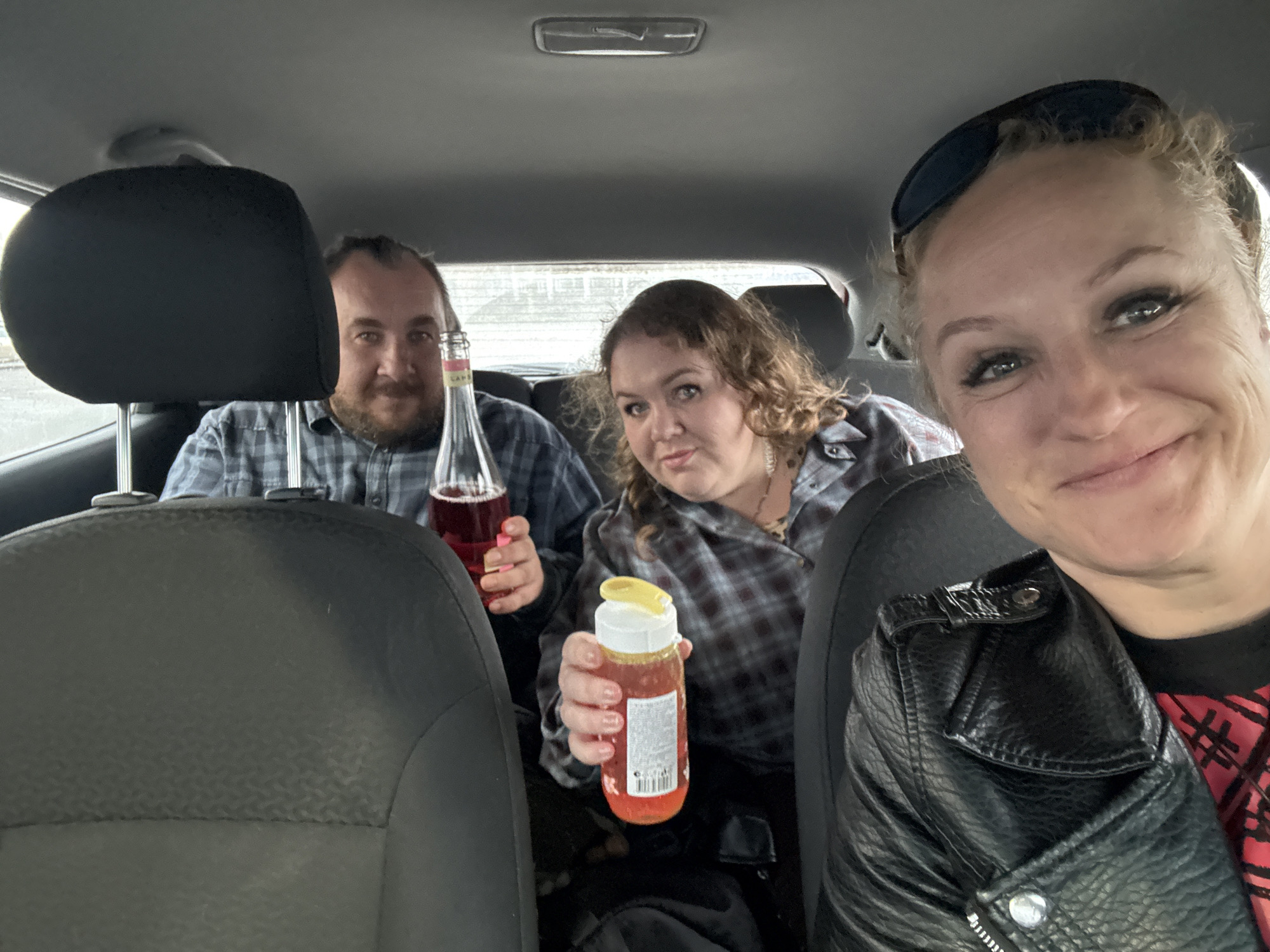 Three people are in a car. A man is in the back seat holding a bottle of red liquid. A woman is in the middle, also in the back seat, holding a yellow bottle with liquid. A woman is in the front seat taking a selfie. They are all wearing plaid shirts. The front seat woman also has on a black leather jacket and sunglasses on her head. The location appears to be a car interior, possibly in transit. It is not possible to confirm that the location is in the city of Izhevsk, Russia, from the visual data provided.