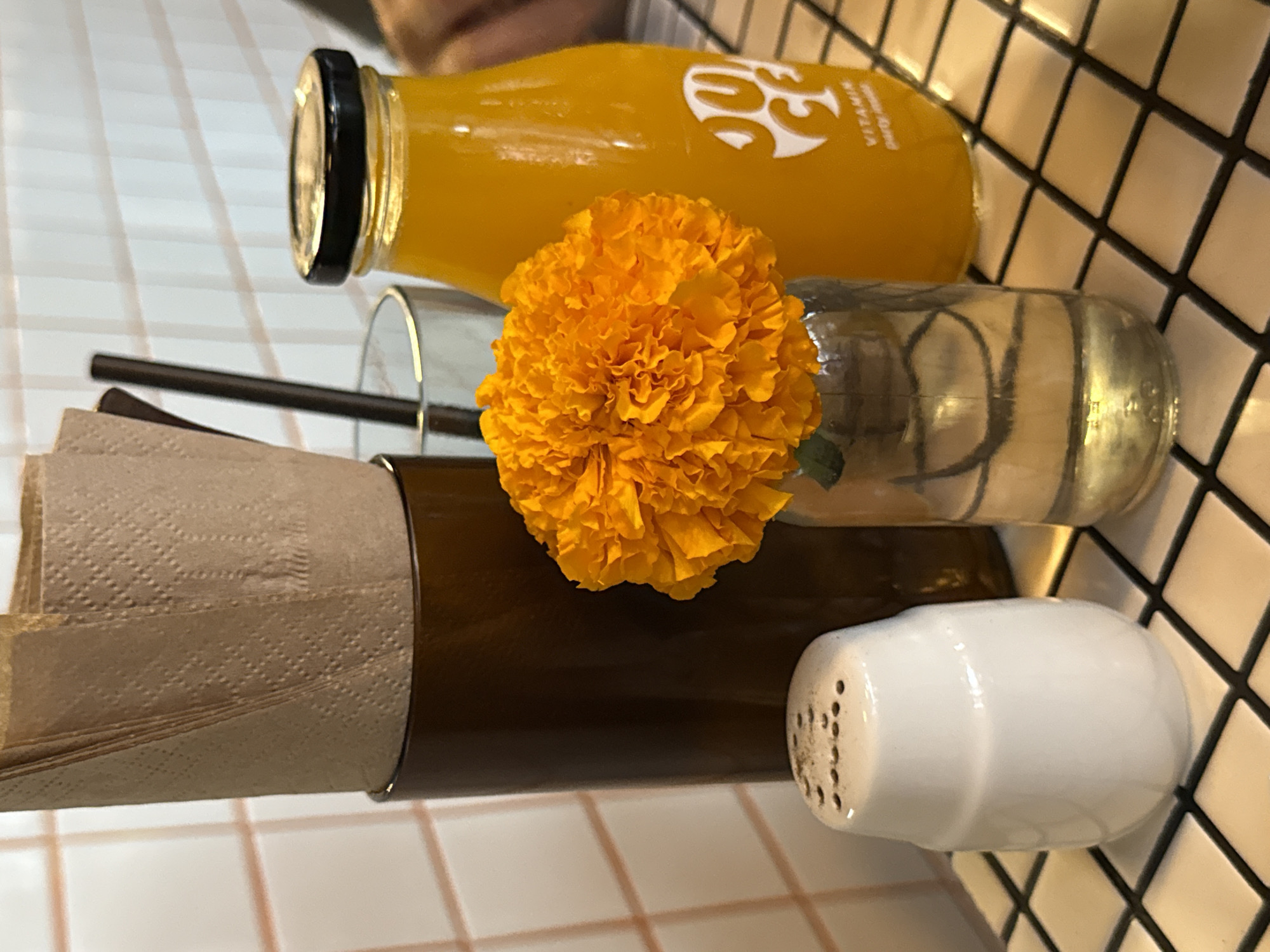 This media file presents an angled, close-up view of a table setting, likely in a cafe or restaurant in North Kuta, Indonesia. The table surface is characterized by white square tiles with contrasting dark grout lines, creating a grid pattern.
In the foreground, a clear glass bottle filled with bright orange juice is prominently displayed, topped with a black screw cap. A white, stylized logo, which appears to be "VC" or similar, is visible on the bottle, along with the word "VITAMIN" printed vertically. Next to the juice bottle, a small, clear glass vase holds a vibrant, densely petaled orange flower, resembling a marigold.
To the left of the flower and vase, a clear drinking glass contains a dark-colored straw. Adjacent to this is a brown holder, possibly metal or wood, filled with neatly folded brown paper napkins, which have a subtle embossed pattern. A small, white ceramic salt shaker with perforated holes on its top is positioned on the right side of the brown holder. A blurred suggestion of a person's hand is faintly visible in the upper background. The scene suggests a fresh and inviting atmosphere, possibly set for a meal or drink during the daytime.