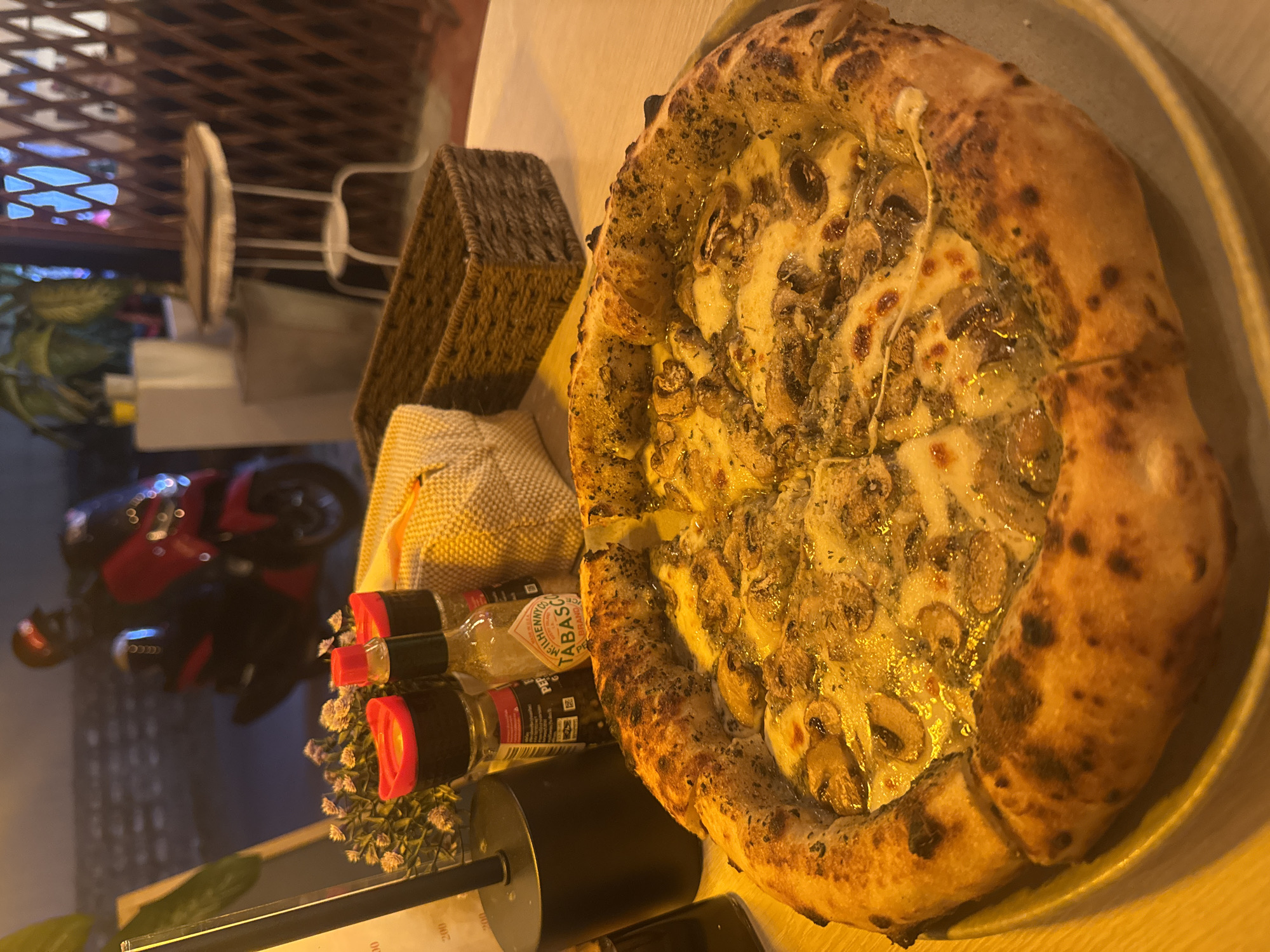 The image shows a close-up of a mushroom pizza on a light-colored plate, likely taken at a restaurant in Baan Mae Nam, Thailand. Beside the plate are condiments, including what appears to be Tabasco sauce, a woven basket, and other containers. In the blurred background, outside the restaurant, a parked red motorcycle is visible, as well as a building with white-framed windows. The lighting suggests it is either dusk or evening.