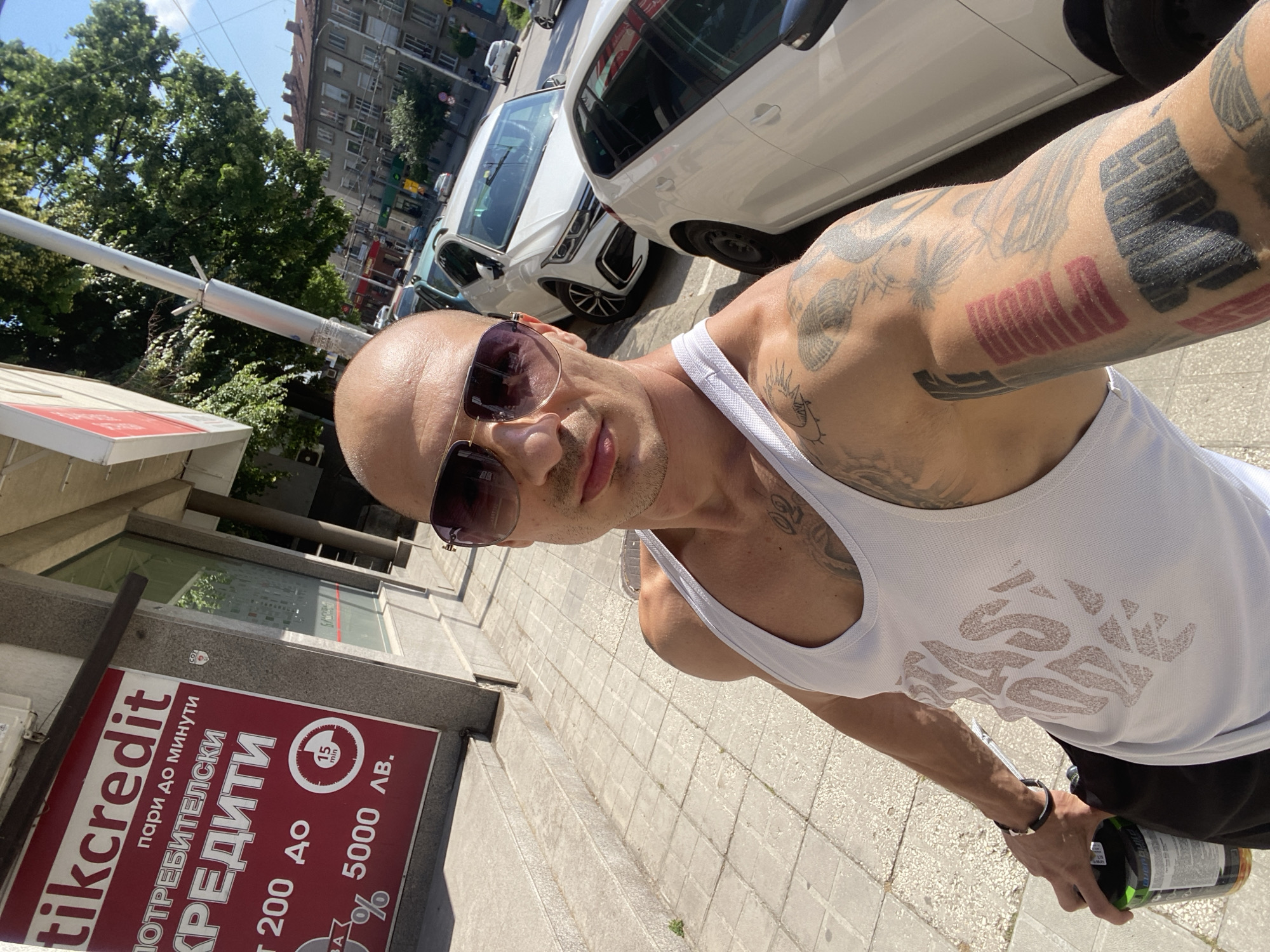 A bald man wearing sunglasses and a white tank top takes a selfie on a sunny day on an urban street, likely in Pleven, Bulgaria. His arms are extensively tattooed with intricate designs, including bold black and red block patterns on his right arm. He holds a green bottle in his left hand. The setting appears to be a sidewalk adjacent to a street lined with parked white cars and multi-story buildings. Trees are visible in the background, suggesting a degree of urban greenery.
The bright sunlight and clear sky indicate it is daytime. On the left, a prominent red and white sign advertises "tikcredit" consumer loans. Visible text on the sign, primarily in Cyrillic, reads: "tikcredit", "пари до минути" (money in minutes), "ПОТРЕБИТЕЛСКИ КРЕДИТИ" (consumer loans), indicating amounts "200 до" (from 200) to "5000 лв." (5000 Bulgarian leva), and "15 min", likely referring to processing time. The man maintains a neutral to slightly smiling and relaxed expression.