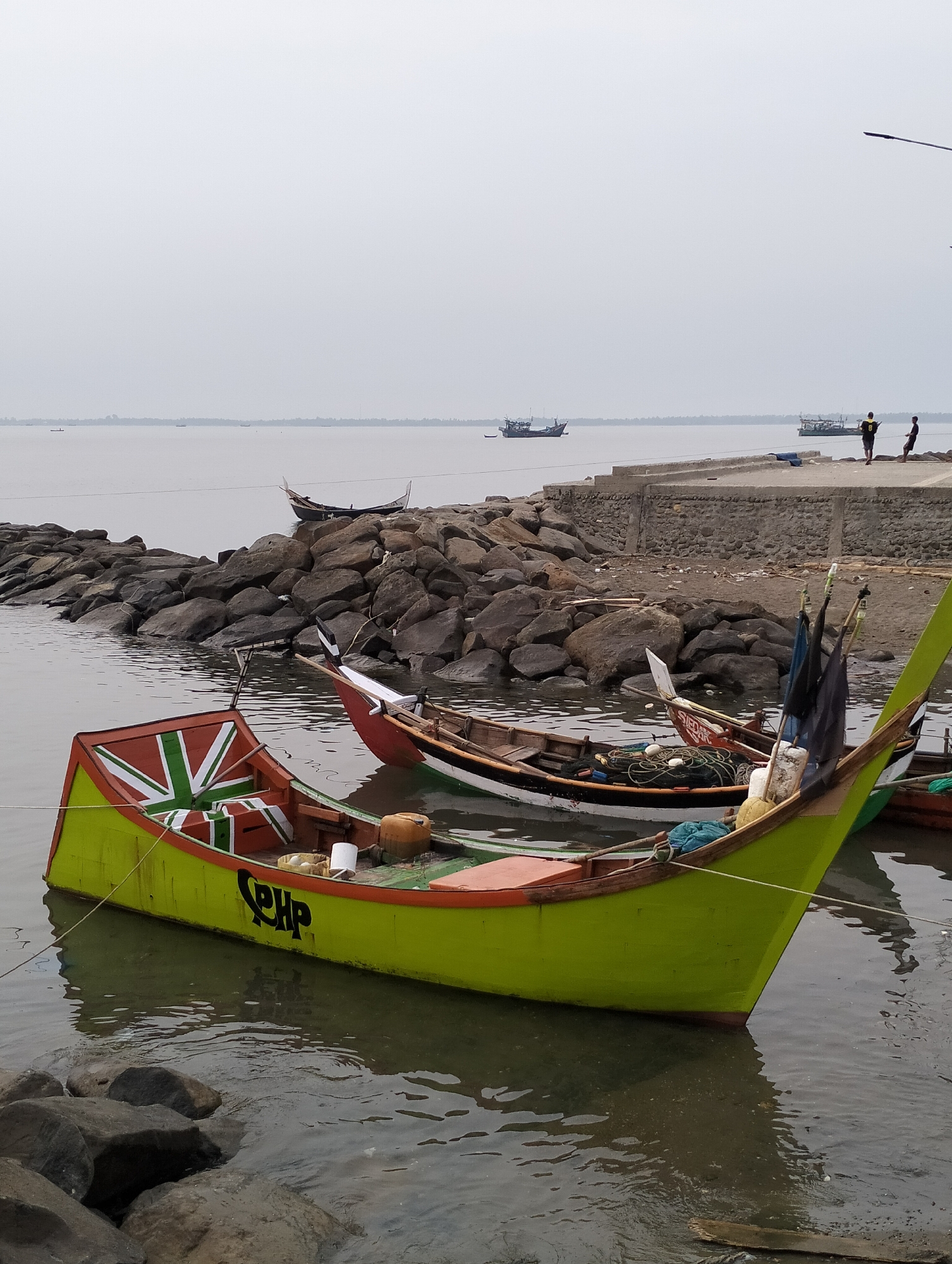 The scene depicts a coastal area in Lhokseumawe, Indonesia, under an overcast, light grey sky. The water in the foreground is calm, greyish-brown, and exhibits minor ripples.
In the immediate foreground, partially submerged, is a wooden fishing boat with a predominantly lime green hull and an orange trim along the gunwale. The boat's bow is painted with a distinctive red, white, and green pattern resembling a Union Jack flag. The side of the hull displays the black letters "SPHP". This boat is tethered by light-colored ropes.
Behind the foreground boat, a rock breakwater, composed of numerous dark grey, irregularly shaped boulders, extends from the right towards the left midground, separating the immediate water from a further section of the coastline.
To the right of the breakwater, a second wooden fishing boat is moored. This vessel features a red upper hull, a white stripe, and a dark lower hull. Fishing nets and ropes are visible within its interior. Two black flags or banners are attached to vertical poles near its bow. Behind the breakwater, a third, dark-hulled wooden boat with a pointed bow is also visible on the water.
On a raised concrete platform or pier to the right, beyond the breakwater, two adult individuals stand with their backs to the viewer. One is wearing a yellow shirt, and the other a darker garment. They appear to be observing the water and the distant horizon. The concrete platform has a textured, possibly stone-faced, side wall.
In the far background, across the expanse of water, a low landmass or coastline is discernible. Several larger vessels, possibly fishing trawlers or cargo ships, are visible on the horizon line.