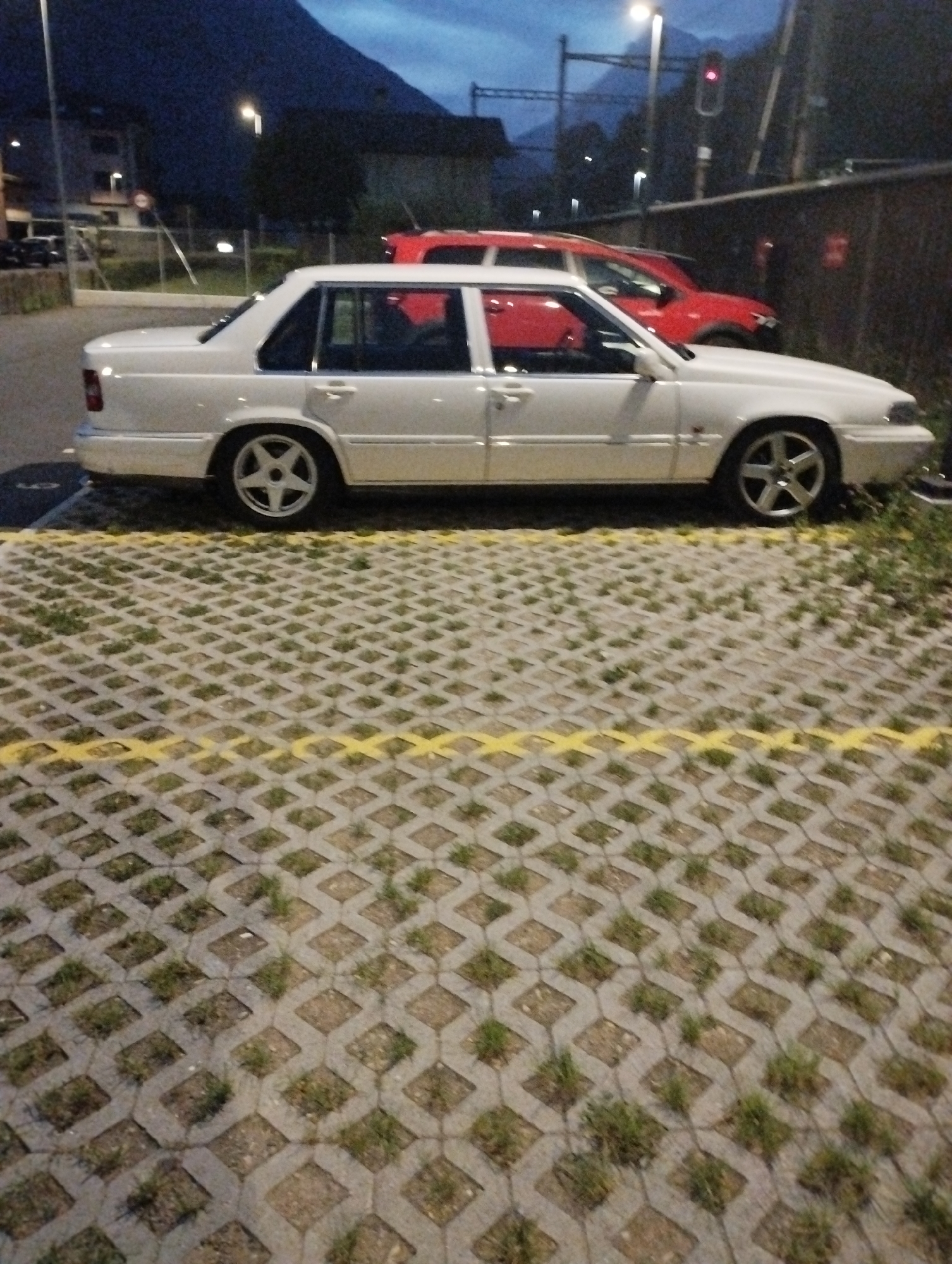 A white Volvo sedan is parked in a parking lot at dusk. The car is facing left and is in the foreground of the image. The parking lot is made of interlocking concrete pavers with grass growing in some of the openings. A yellow line is painted across the parking lot. In the background, there is a red SUV, a traffic light, trees, buildings, and a mountain. The sky is dark blue, indicating it is either early morning or late evening.