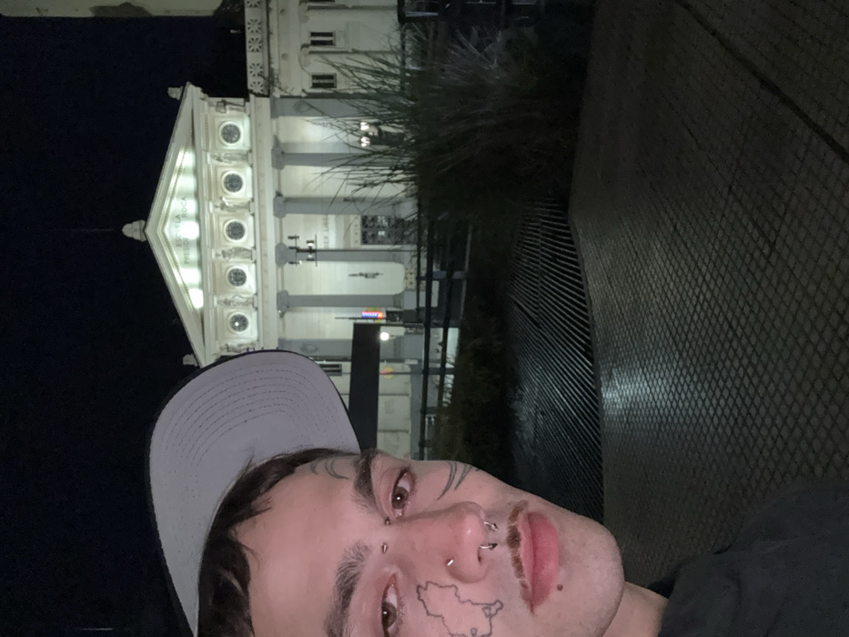 A low-angle, tilted shot features a person with facial piercings and tattoos wearing a baseball cap. Behind the person is a brightly lit, ornate building with classical columns and windows, possibly a library. To the right of the building is a blurred arrangement of foliage and a geometric structure. The scene appears to be at night.