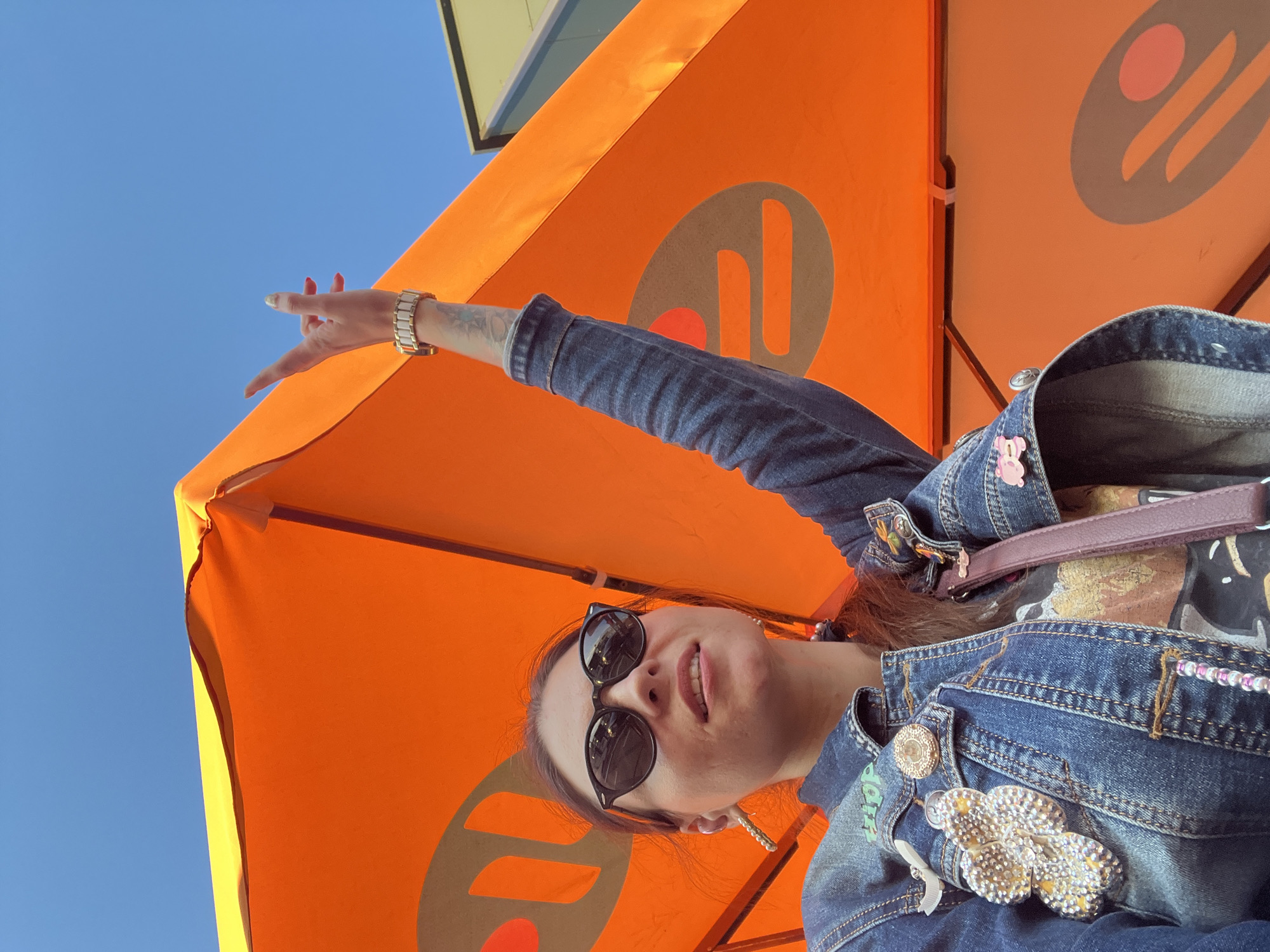 A low-angle medium shot captures a woman from the chest up, standing outdoors under an orange umbrella with a blue sky in the background. The woman, who appears to be Caucasian with brown hair, is wearing sunglasses, a denim jacket adorned with various patches and pins, and a T-shirt. She is looking up and to her left, with her right arm raised, her index and pinky fingers extended in a gesture resembling the "rock on" sign. A prominent watch is visible on her wrist. The umbrella has a repeating circular logo with orange and grey elements. The scene is devoid of any specific landmarks that would indicate a location in Kostroma, Russia, or any other city.